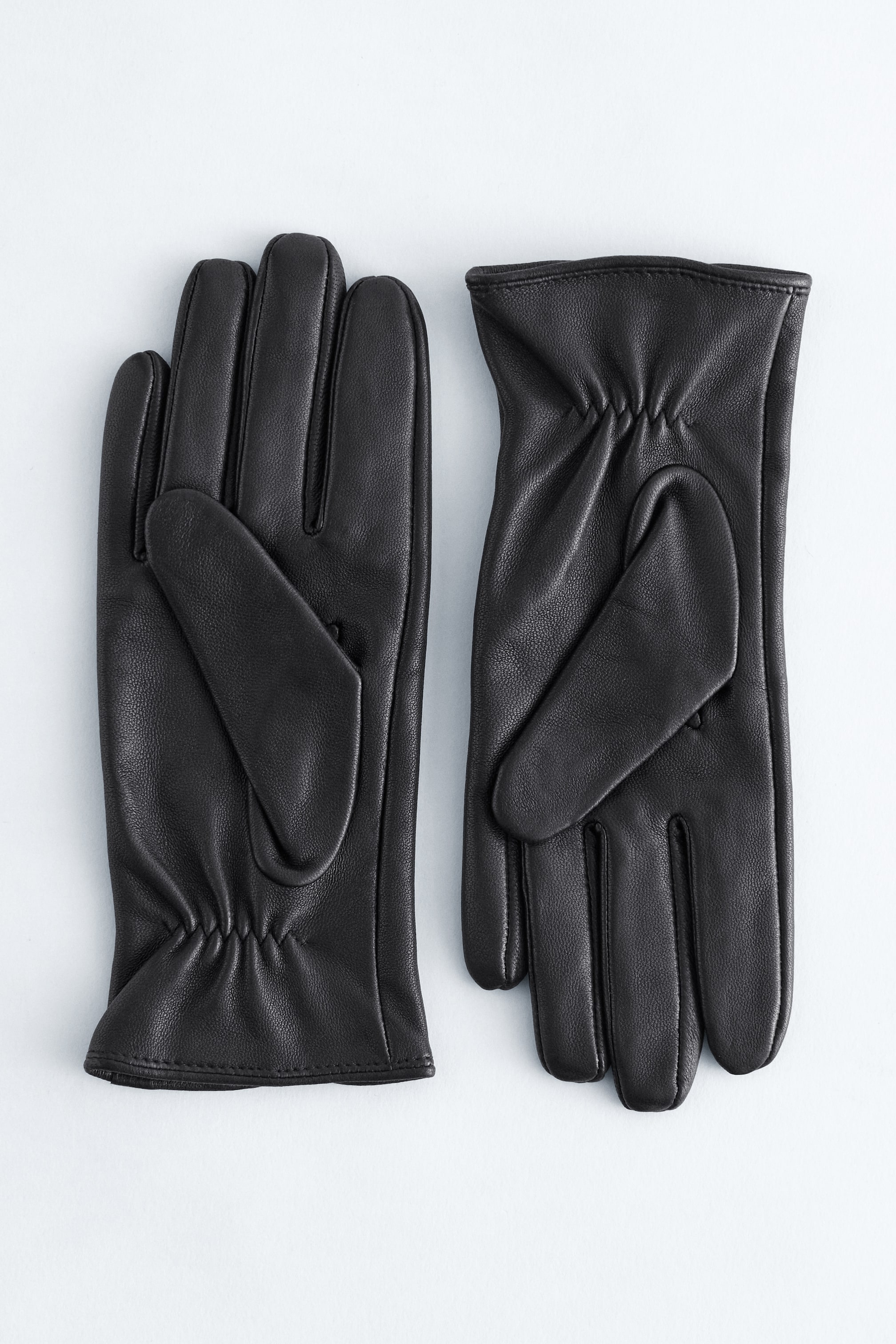 View larger image: A pair of black leather gloves is lying flat. Each glove is featuring a smooth finish, a classic slit on the back of the wrist, and elasticized gathering on the palm side of the wrist.