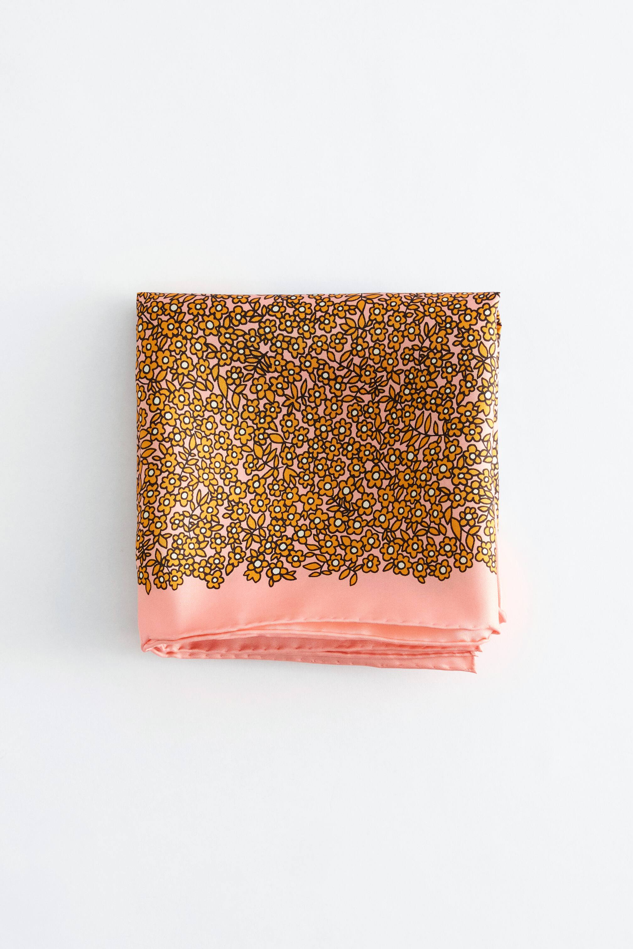 Visualizza immagine più grande: Una sciarpa è piegata a forma di quadrato, mostrando una base rosa. Presenta un fitto motivo di piccoli fiori giallo-arancio con centri bianchi e foglie, tutti delineati in nero. Un bordo rosa solido è visibile lungo il bordo inferiore.