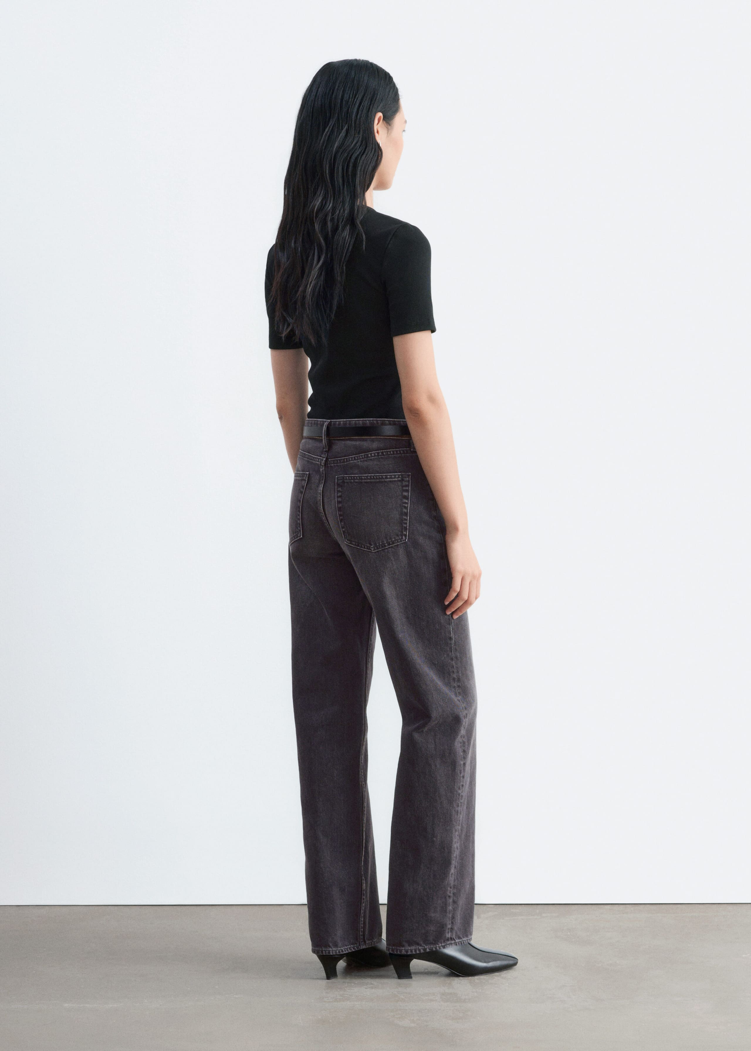 A black, ribbed short-sleeve T-shirt is being worn by the model, tucked into a pair of dark grey jeans. The slim-fit T-shirt is shown from a rear three-quarter angle, highlighting its textured finish and clean silhouette.