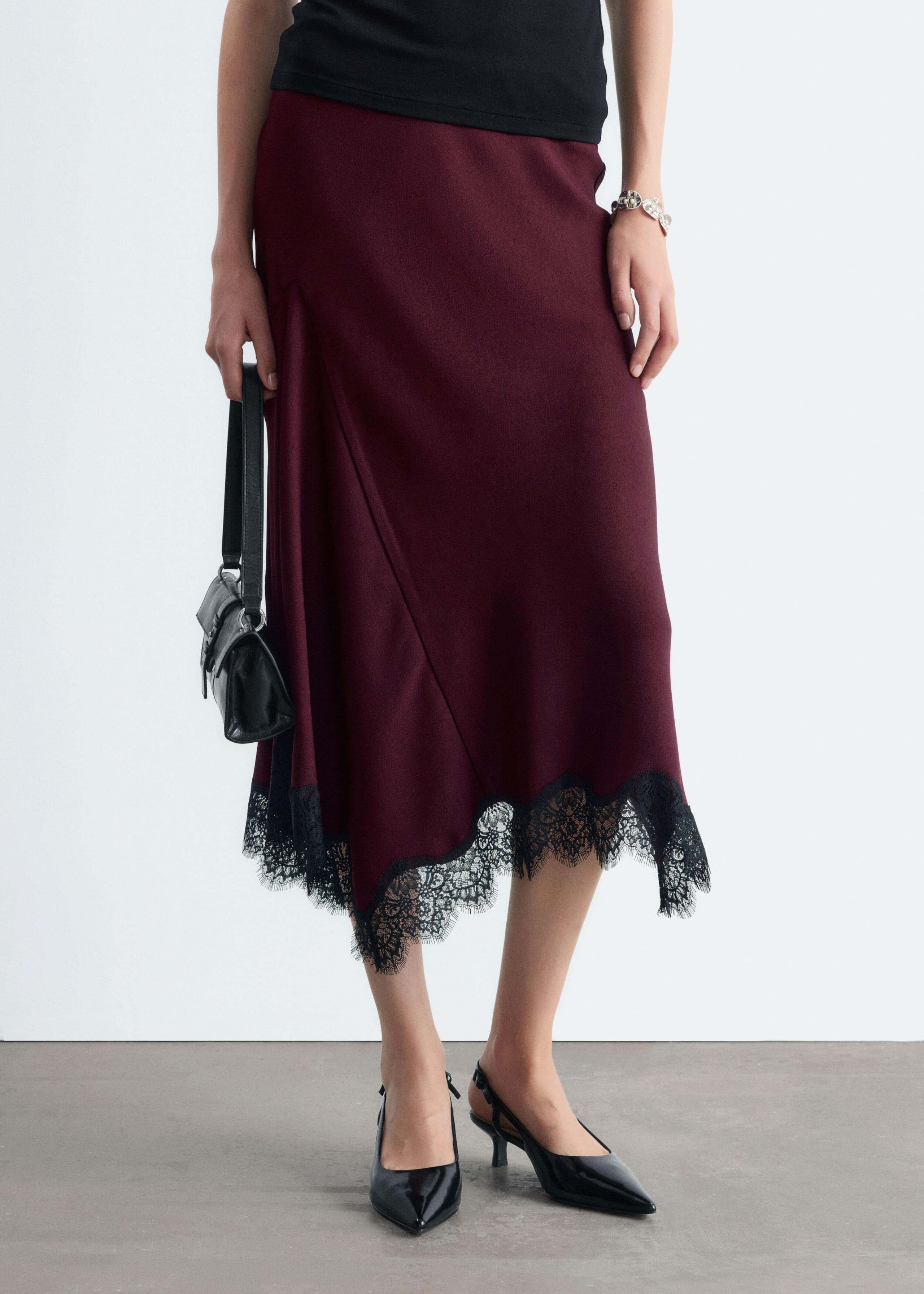 A burgundy flared midi skirt with an asymmetrical hemline trimmed with delicate black lace is worn by the model. The skirt is styled with a black top, black slingback heels, and a small black handbag carried in one hand.