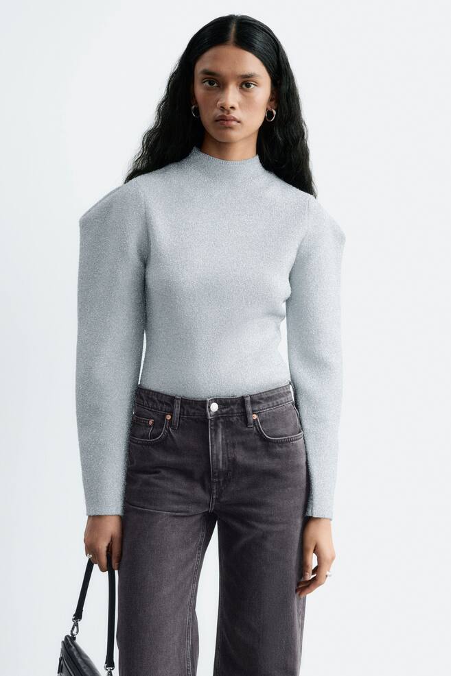 De model draagt een lichtblauwe, glinsterende gebreide top met een hoge hals en gestructureerde, geplooide schouders. De nauwsluitende top is ingestopt in donkergrijze jeans. - 1