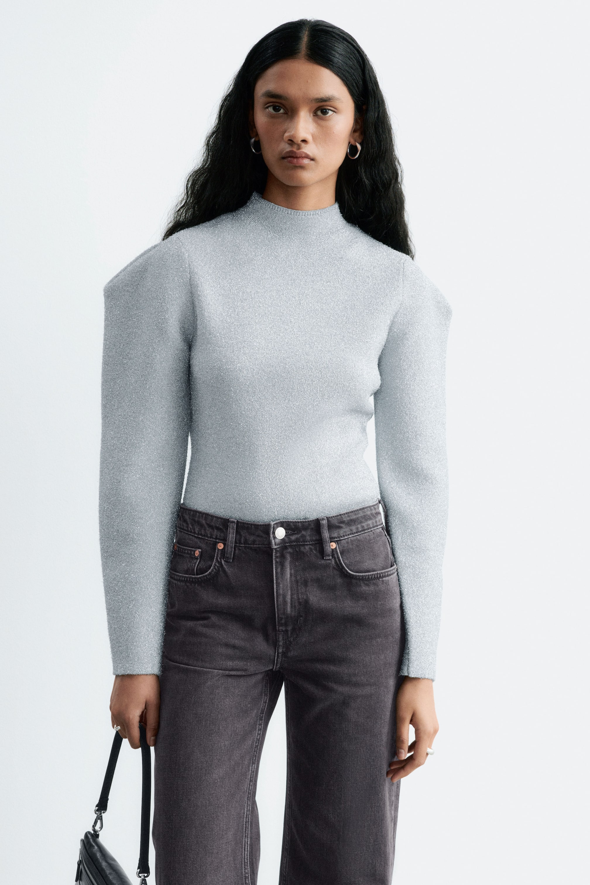 Ver imagen más grande: La modelo lleva un top de punto azul claro con brillo, con cuello alto y hombros estructurados con pliegues. El top de corte entallado está metido por dentro de unos vaqueros de color gris oscuro.