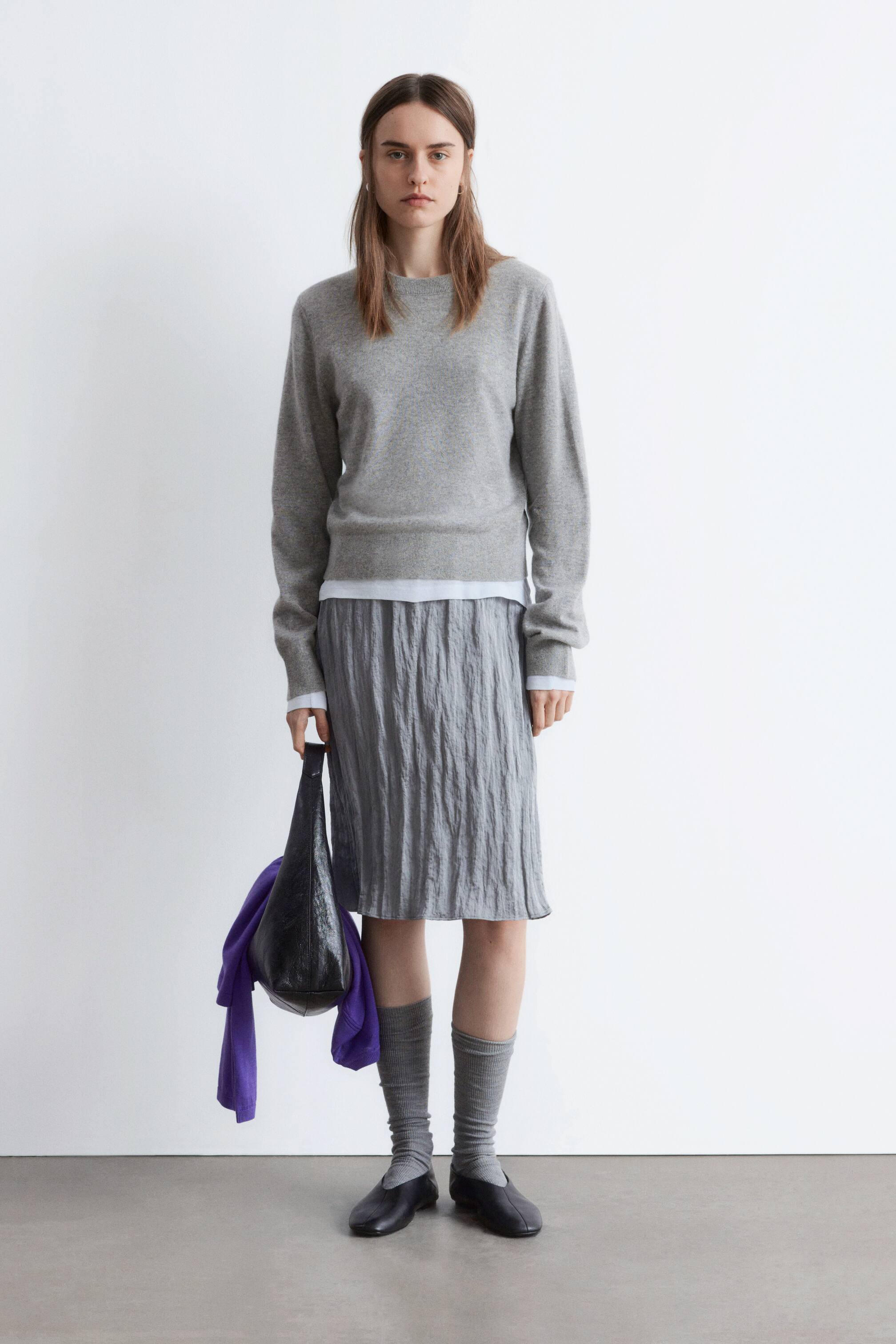 Größeres Bild ansehen: Ein grauer Pullover mit Rundhalsausschnitt, geripptem Saum und Bündchen wird über einem weißen langärmeligen Hemd getragen und mit einem grauen knielangen, geknitterten Rock kombiniert. Graue gerippte Socken, die bis zur Mitte der Wade reichen, und schwarze flache Slipper werden getragen, und eine schwarze Hobo-Tasche mit einem lila drapierten Kleidungsstück wird getragen.