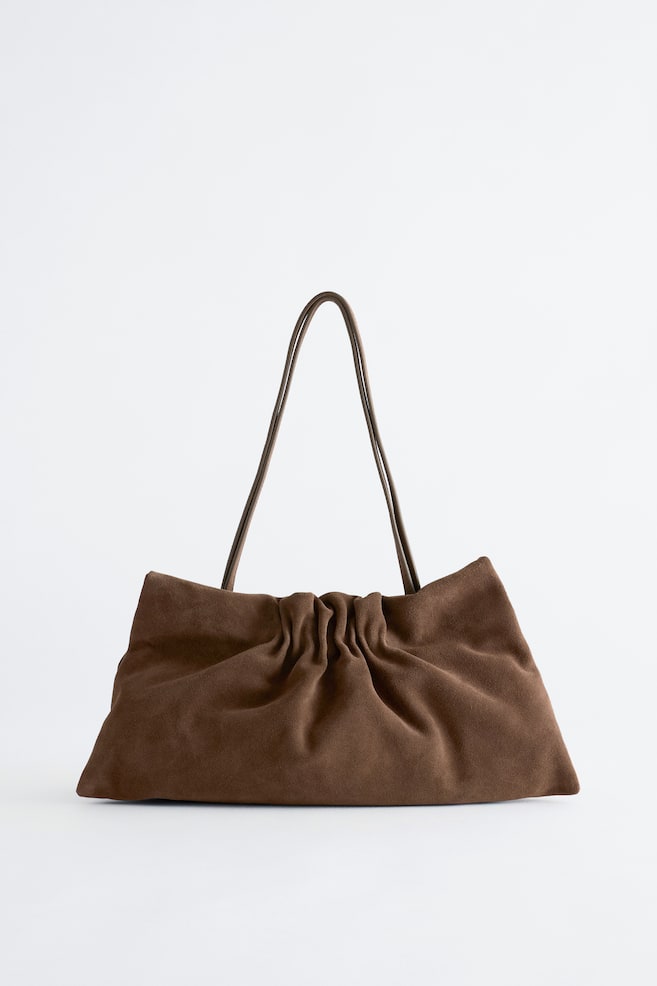 Un sac à bandoulière en daim marron souple est présenté de face, avec une silhouette rectangulaire souple et décontractée, rehaussée de détails froncés et plissés au centre. De fines bretelles assorties en cuir sâélèvent depuis le haut. - 1