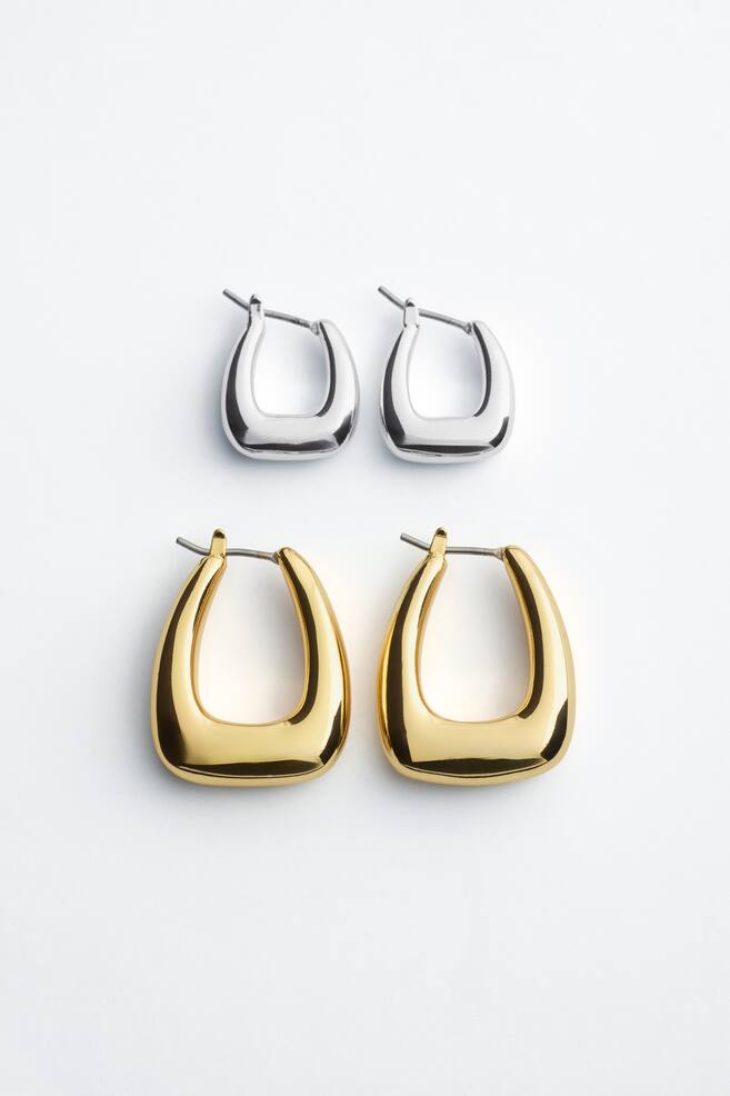 Two pairs of sculptural, chunky hoop earrings are lying flat, displaying a smaller silver-toned pair positioned above a larger gold-toned pair. Both pairs feature a squared U-shape with a polished finish and hinge-post closures. - 1