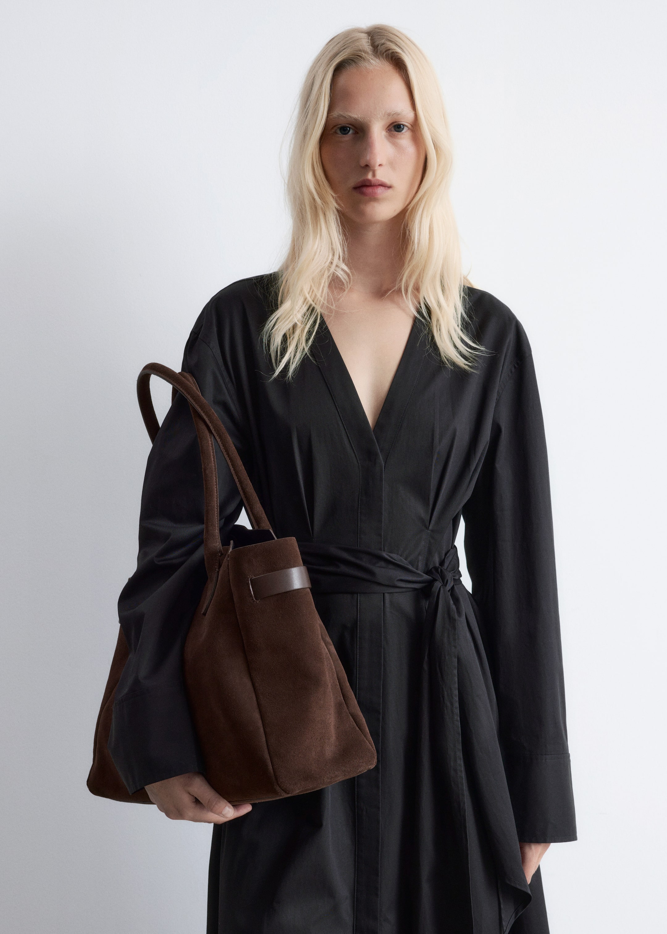 Visualizza immagine più grande: Una borsa tote in camoscio marrone viene tenuta, mostrando la sua forma morbida e non strutturata. Due manici arrotondati superiori e un dettaglio di cinturino orizzontale sono visibili sulla parte superiore anteriore, mentre è inclinata leggermente verso sinistra.