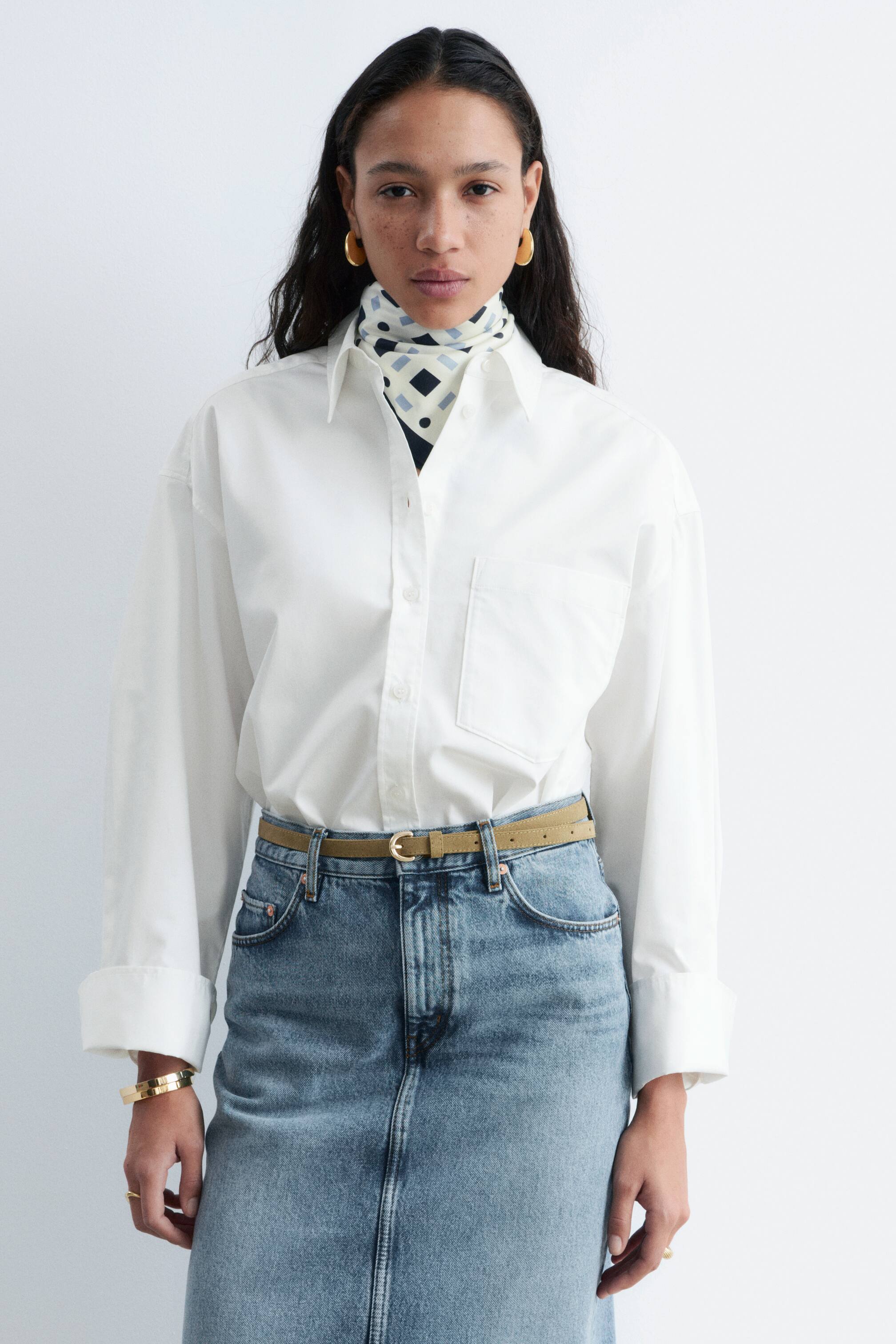 Nagyobb kép megtekintése: Egy fehér, gombos ing van bemutatva, amely hegyes gallérral, elülső gombsorral és egyetlen foltzsebbel rendelkezik a bal mellkason. Hosszú ujjai a mandzsettáknál fel vannak tűrve, és egy farmer szoknyába betűrve viselik, a nyitott gallér alatt pedig egy mintás sál látható.