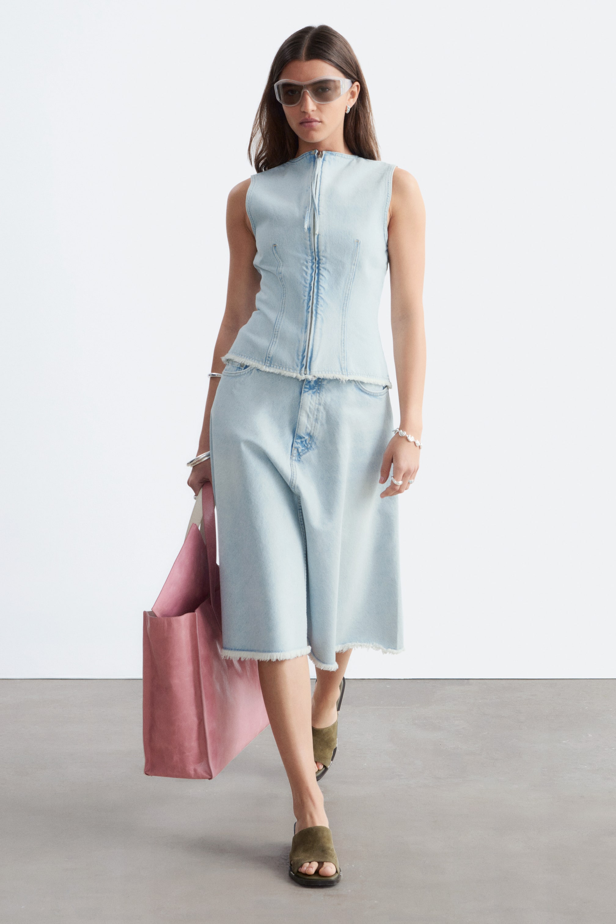 Visualizza l'immagine ingrandita: L'abito due pezzi in denim azzurro chiaro presenta un top senza maniche con colletto alto, chiusura a zip frontale e corpetto con cuciture, il tutto rifinito con un orlo sfrangiato. La gonna midi a trapezio coordinata presenta anch'essa un orlo sfrangiato, mostrata da una vista frontale leggermente angolata.