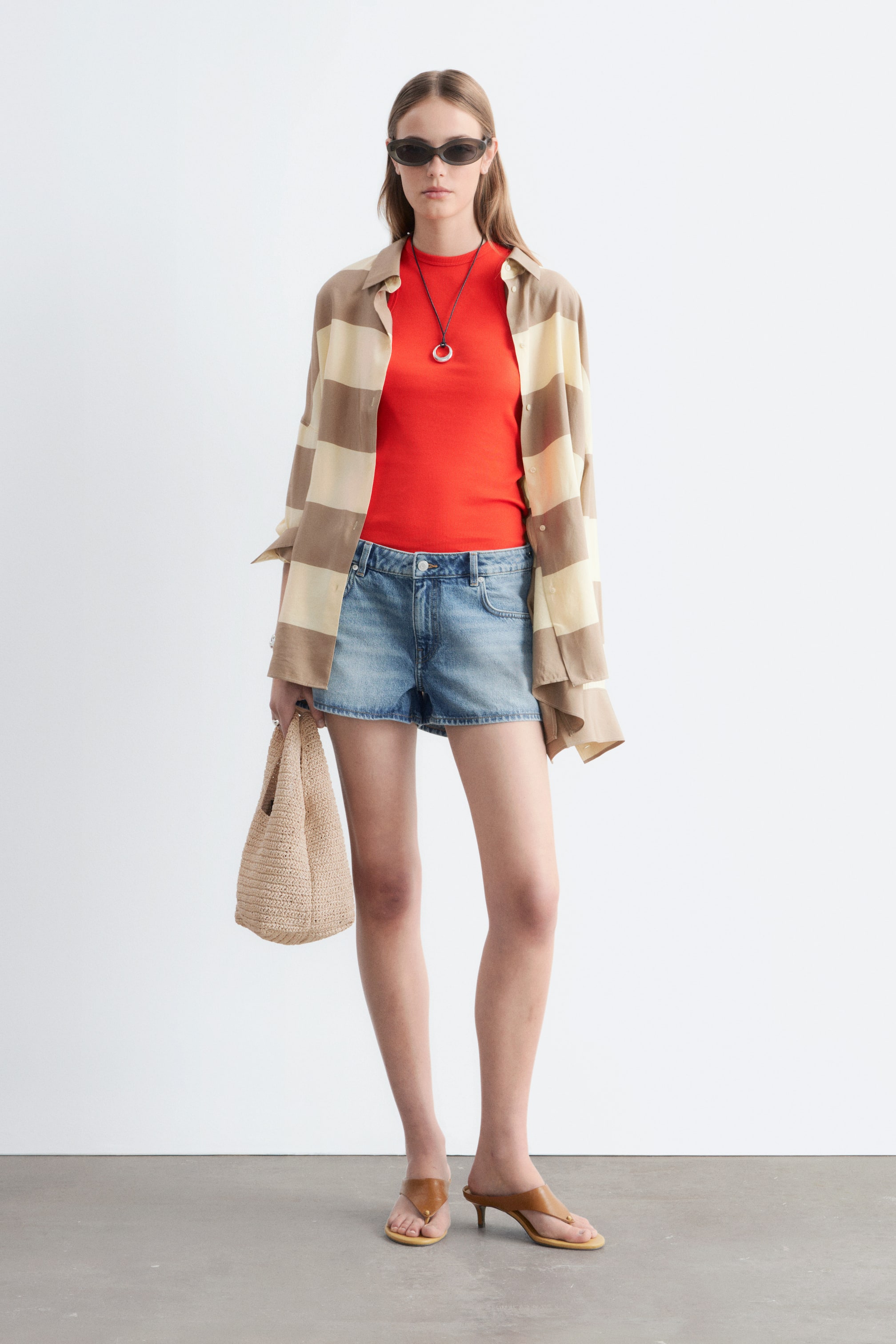 Agrandir l'image: Un haut rouge ajusté est superposé sous une chemise ouverte à rayures beige et crème, assorti à un short en denim délavé clair avec un ourlet effiloché. Des sandales à talon kitten marron à lanières, des lunettes de soleil ovales foncées, un collier en corde noire avec un pendentif circulaire, et un petit sac fourre-tout en paille tressée complètent l'ensemble.