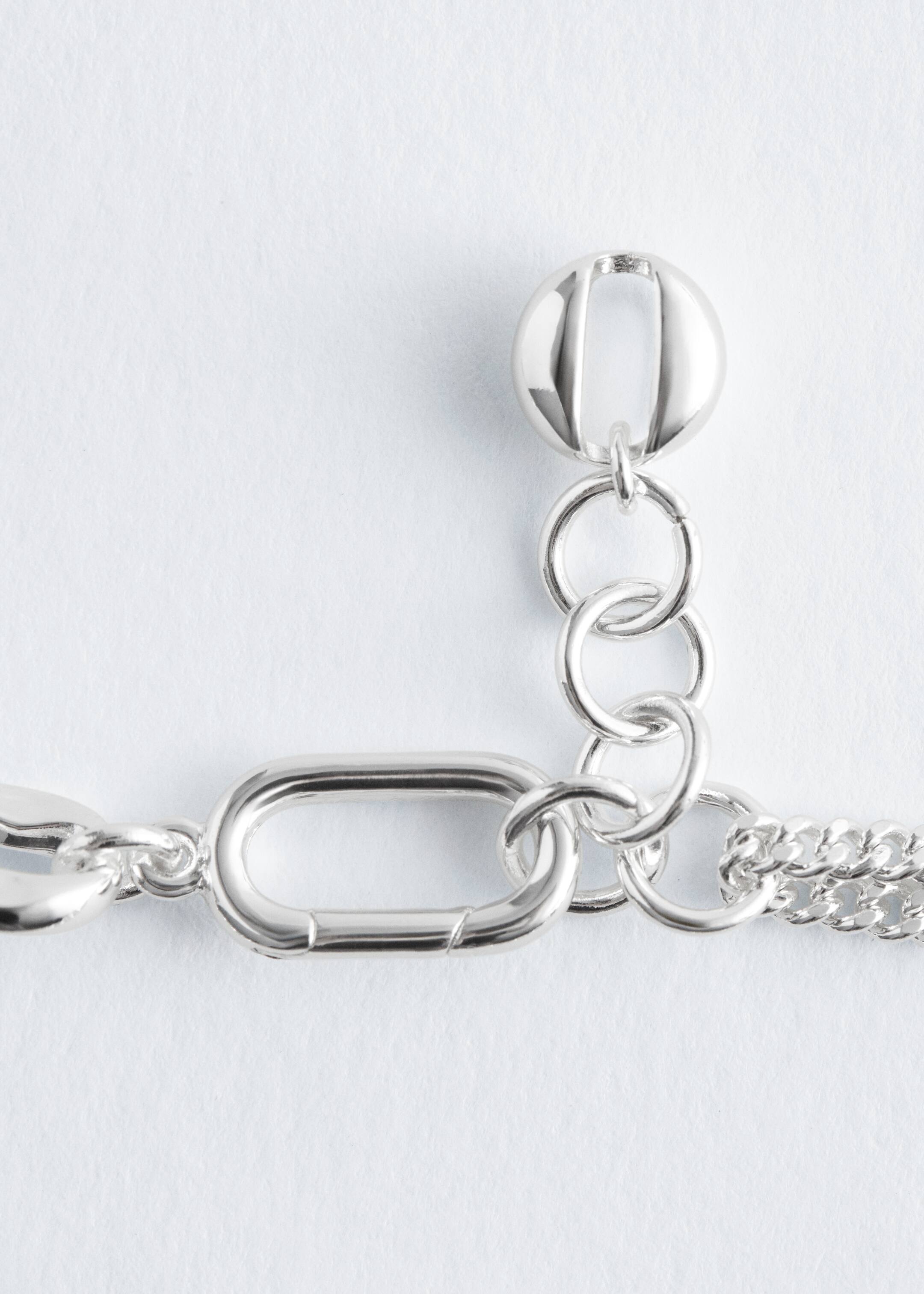 View larger image: Necklace, BACK. A silver-toned necklace is featuring an elongated, rounded O-ring clasp, with an extension chain of smaller round links terminating in a spherical end piece. A section of a curb chain is also visible.