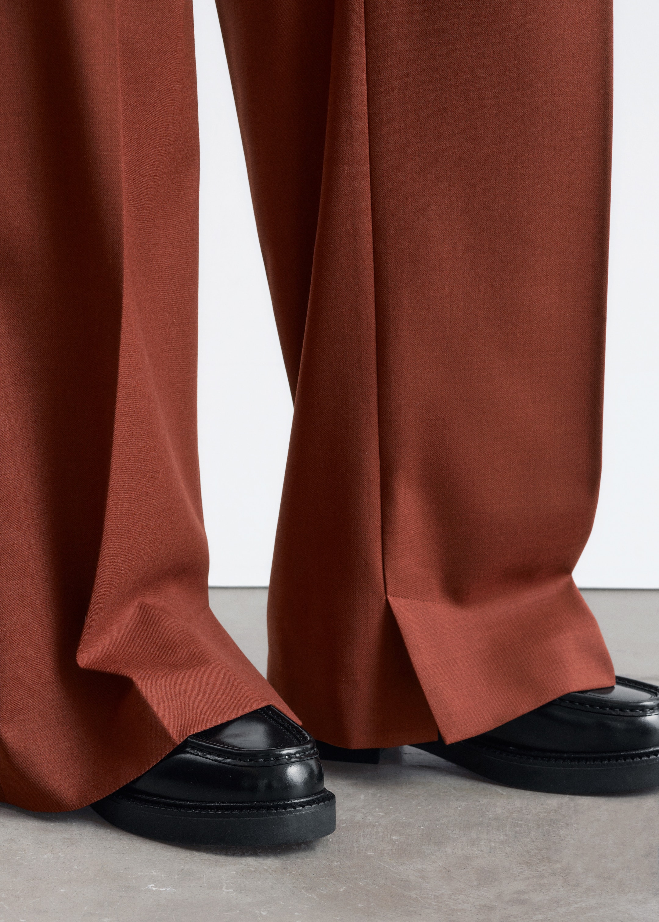 Größeres Bild ansehen: Rostfarbene, weit geschnittene Hosen fließen über schwarze Mokassin-ähnliche Loafer mit dicken Sohlen, aus einem niedrigen, frontalen Winkel betrachtet, der ihre entspannte Passform und lange Länge betont. Die Hosen haben eine subtile vordere Falte am linken Bein und sind mit kurzen Knöchelschlitzen an den äußeren Säumen gestaltet.