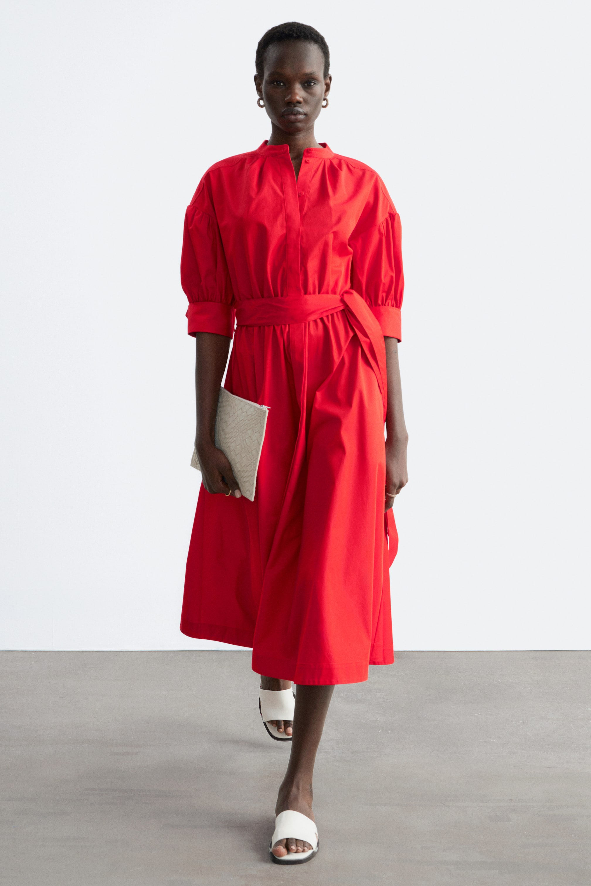 Ver imagem maior: Um vestido midi vermelho, com gola de banda, carcela de botões na frente e mangas bufantes a três quartos com punhos, está a ser usado pela modelo. Um cinto de tecido a condizer está amarrado ao lado, e a modelo segura uma clutch de palha de cor clara enquanto também usa sandálias brancas de enfiar.