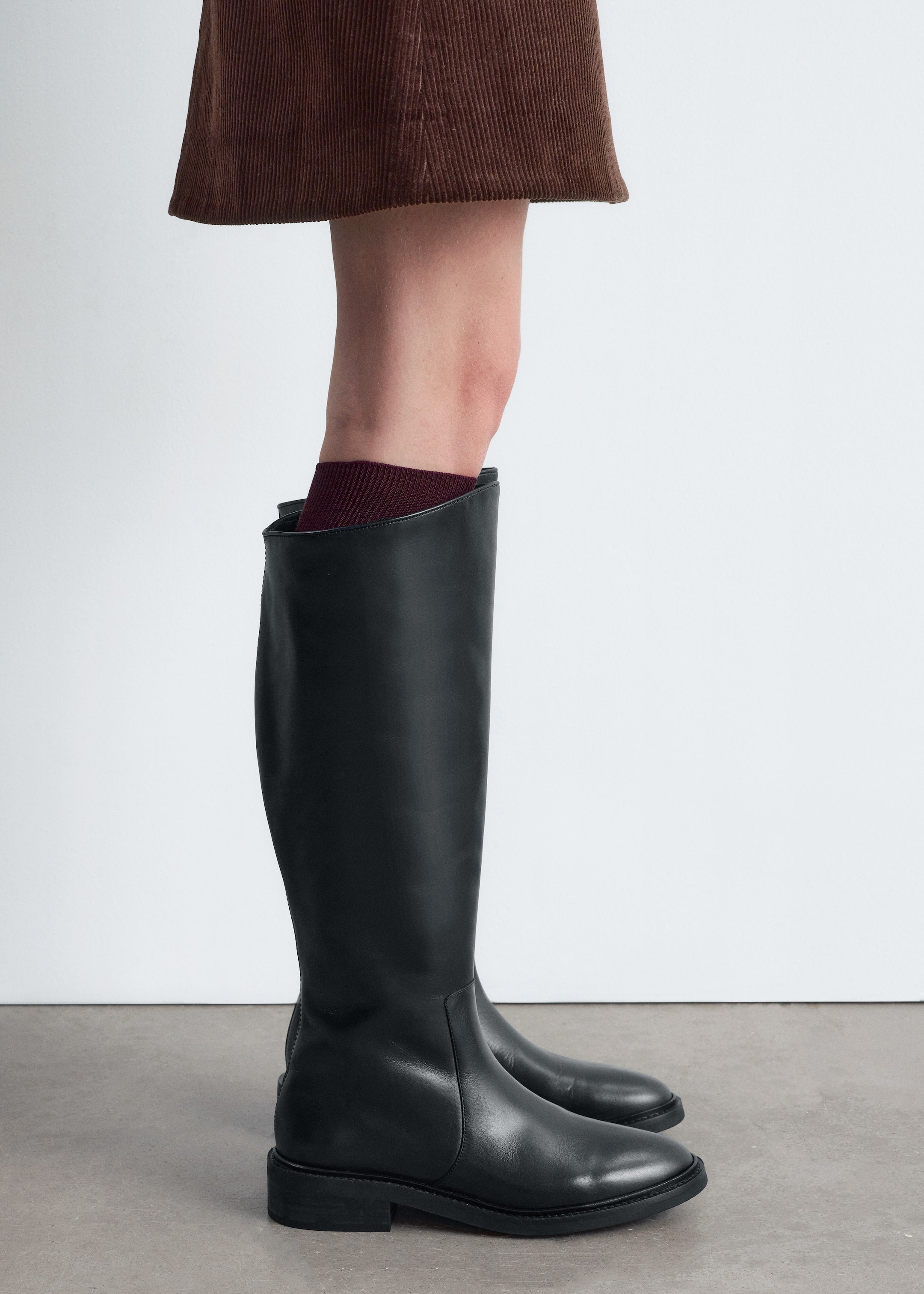 Größeres Bild anzeigen: Ein Paar schwarze, glatte Lederstiefel in Kniehöhe wird im Seitenprofil präsentiert, mit einem hohen, strukturierten Schaft, einer abgerundeten Spitze und einem niedrigen Blockabsatz. Ein bordeauxroter gerippter Socken ist am Stiefelrand sichtbar, mit einem braunen Cordrock, der darüber hängt.