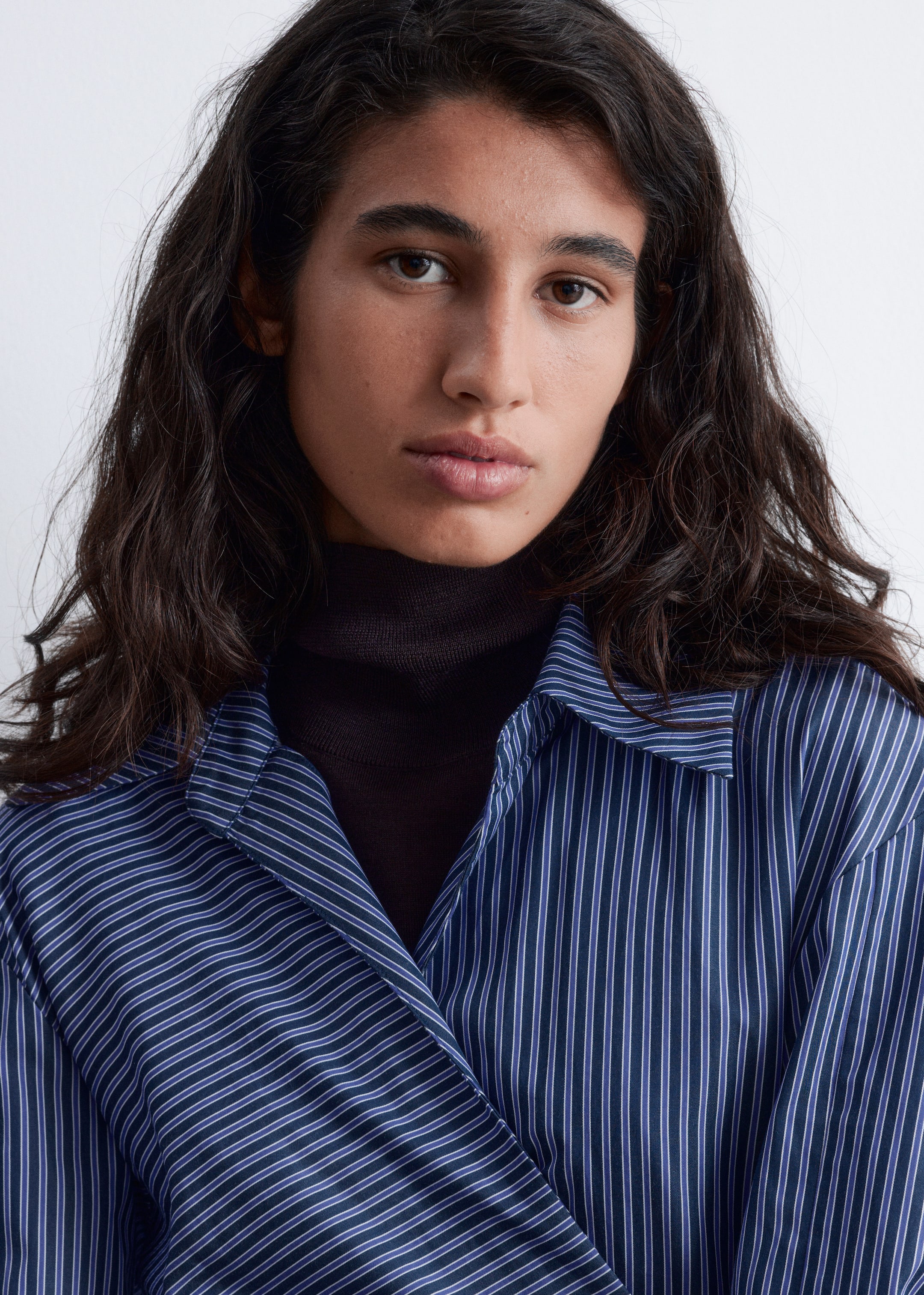 Visualizza immagine più grande: Una camicia blu scuro con sottili strisce verticali bianche e viola è sovrapposta a un dolcevita marrone scuro. Il colletto della camicia è visibile e i suoi pannelli frontali si incrociano sul petto.
