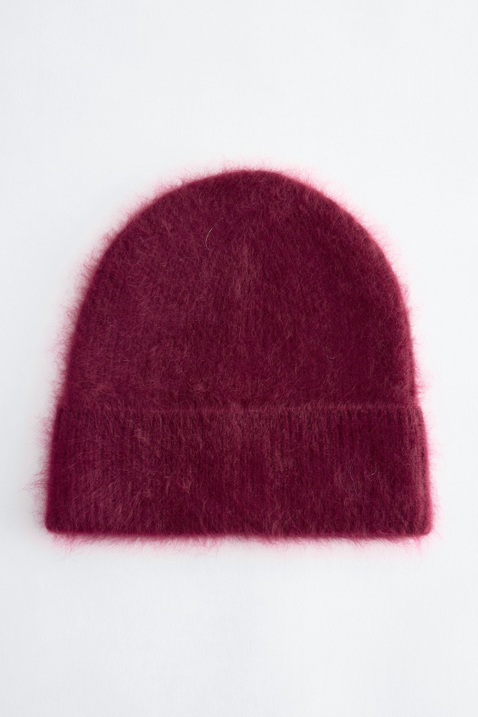 Un cappello beanie in maglia color bordeaux è mostrato frontalmente, mettendo in evidenza una texture soffice e pelosa, con una calotta arrotondata e un risvolto a costine ripiegato.