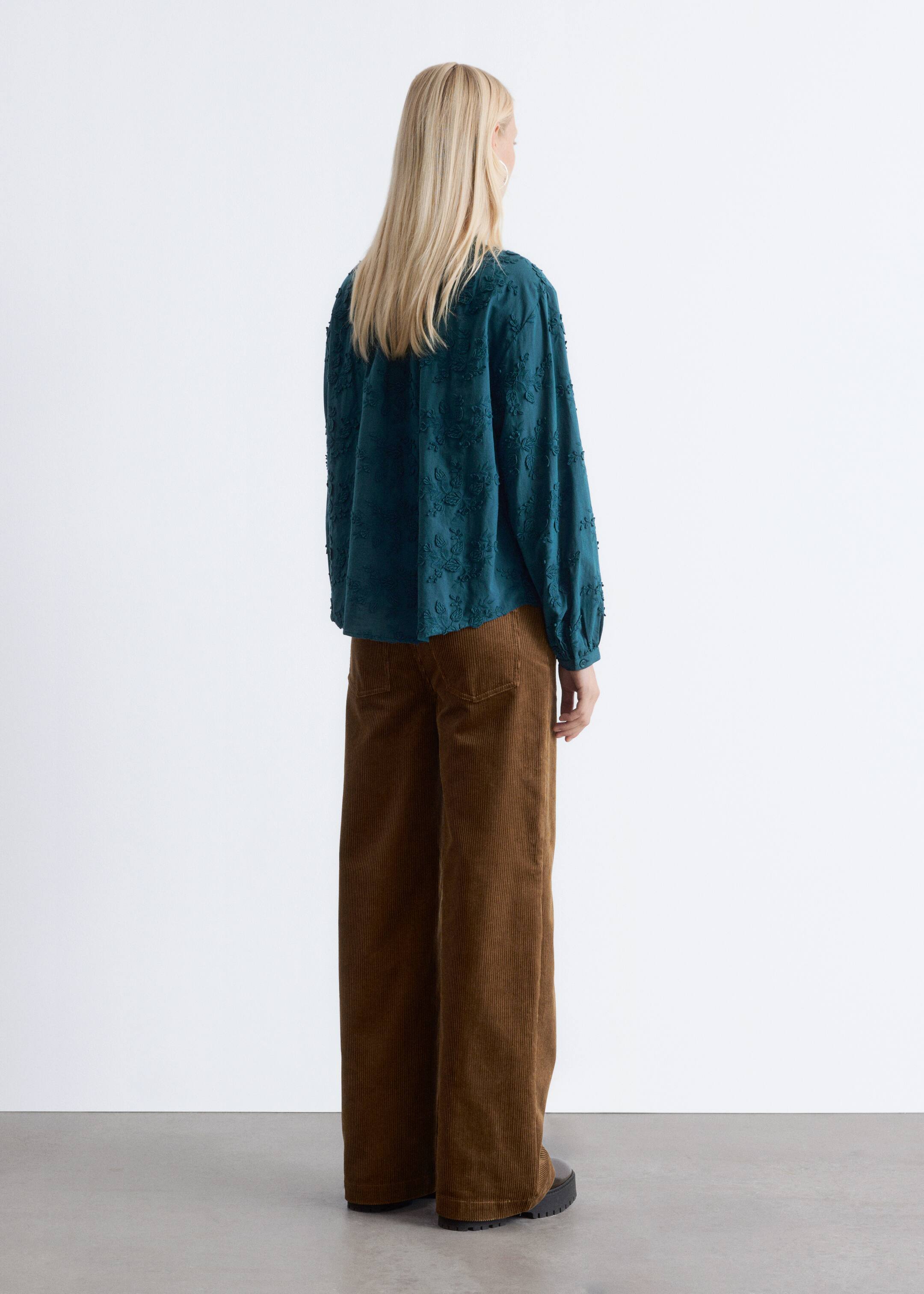Agrandir l'image: Une blouse couleur sarcelle avec une broderie florale texturée et des manches longues froncées est portée avec un pantalon en velours côtelé marron à jambes larges, vue de dos.