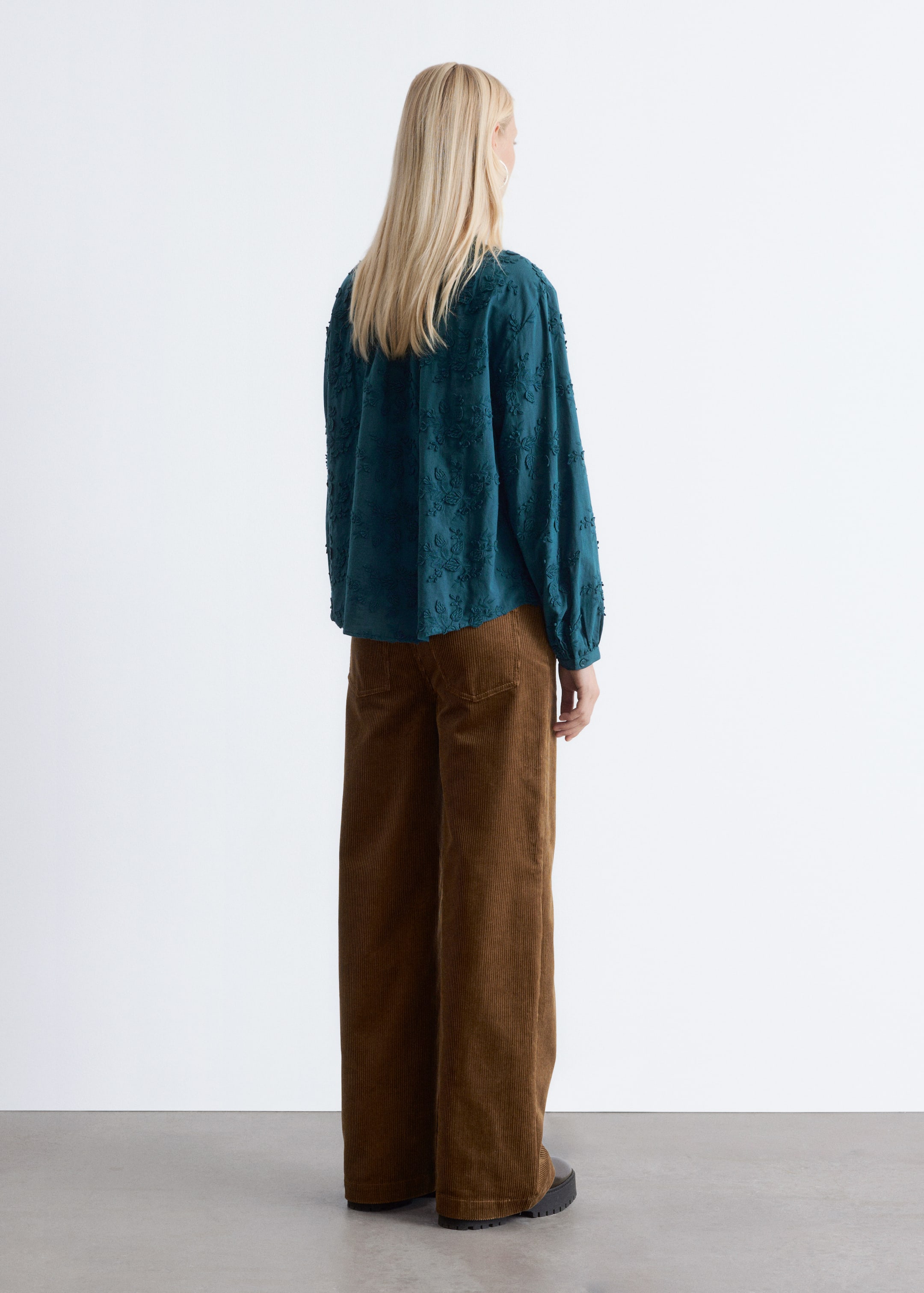 Visualizza immagine più grande: Una blusa color teal con ricami floreali in rilievo e maniche lunghe arricciate è indossata con pantaloni a gamba larga in velluto a coste marrone, vista da dietro.