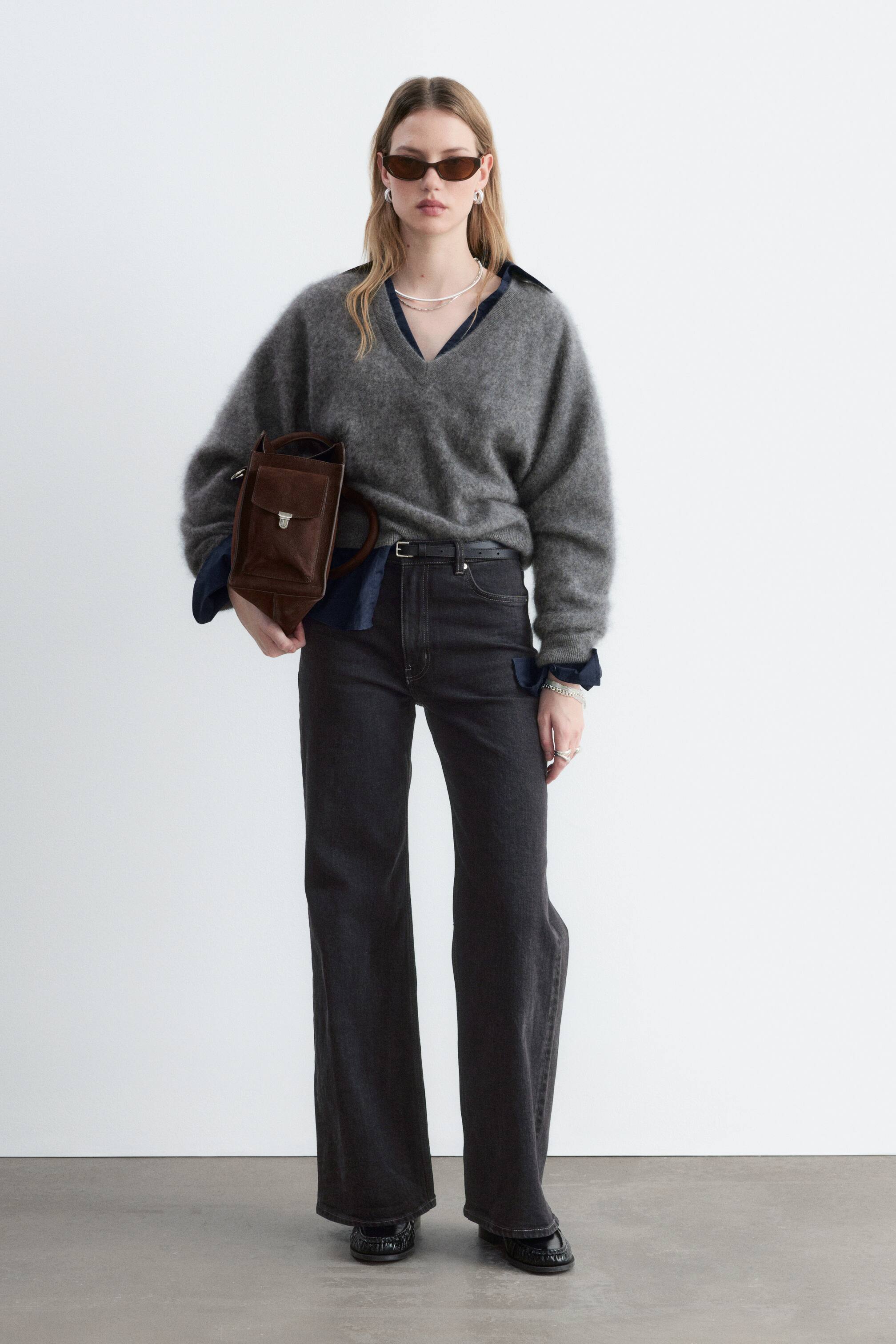 Ver imagen más grande: La modelo lleva unos vaqueros de tiro alto y color gris oscuro, de pernera ancha y ajustados con un cinturón negro fino. Los vaqueros se combinan con un jersey gris de cuello en V sobre una camisa azul con cuello y se complementan con zapatos negros.