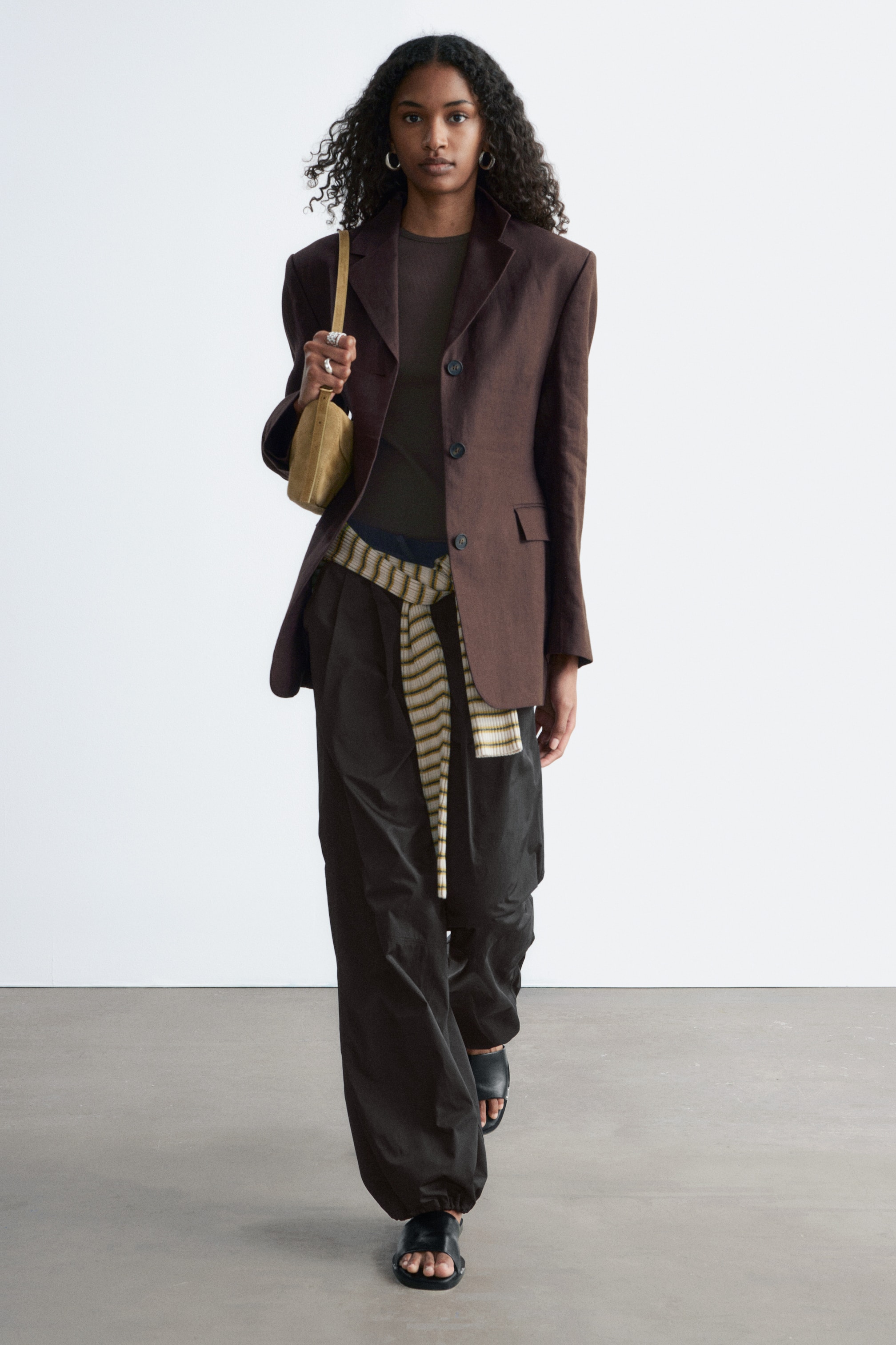 Vis større bilde: Mørke twillbukser bæres av modellen, med en avslappet barrel-leg silhuett med frontplisser, vertikale kuttlinjesømmer og samlet elastisk kant. Et stripete stoff er knyttet rundt livet over buksene.