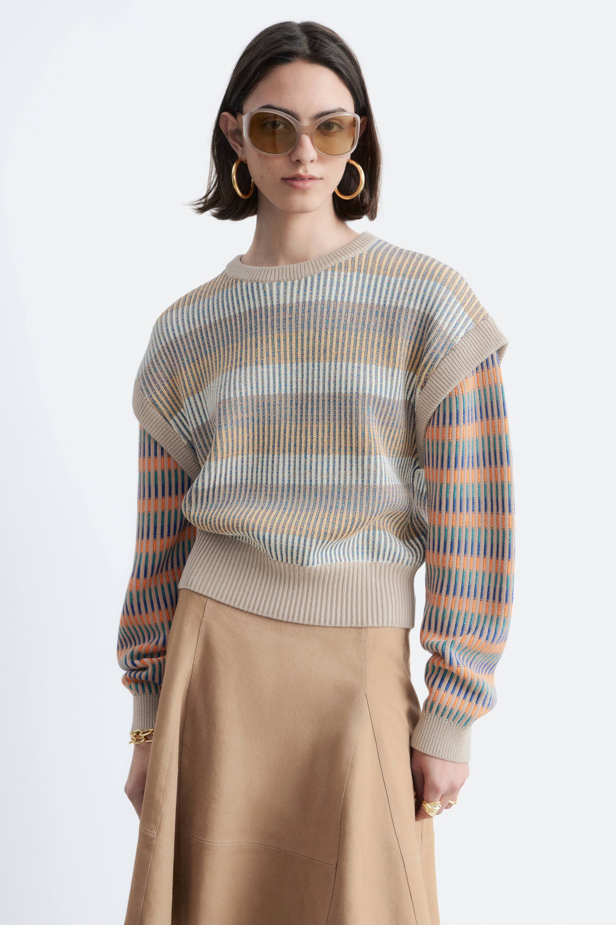 Преглед на по-голямо изображение: Моделът носи скъсен, широк плетен пуловер, който се отличава с оребрена текстура и многоцветни вертикални райета на тялото и ръкавите. Пуловерът има кръгло деколте, спуснати рамене и дълги ръкави с оребрени маншети, и е съчетан с бежова пола.