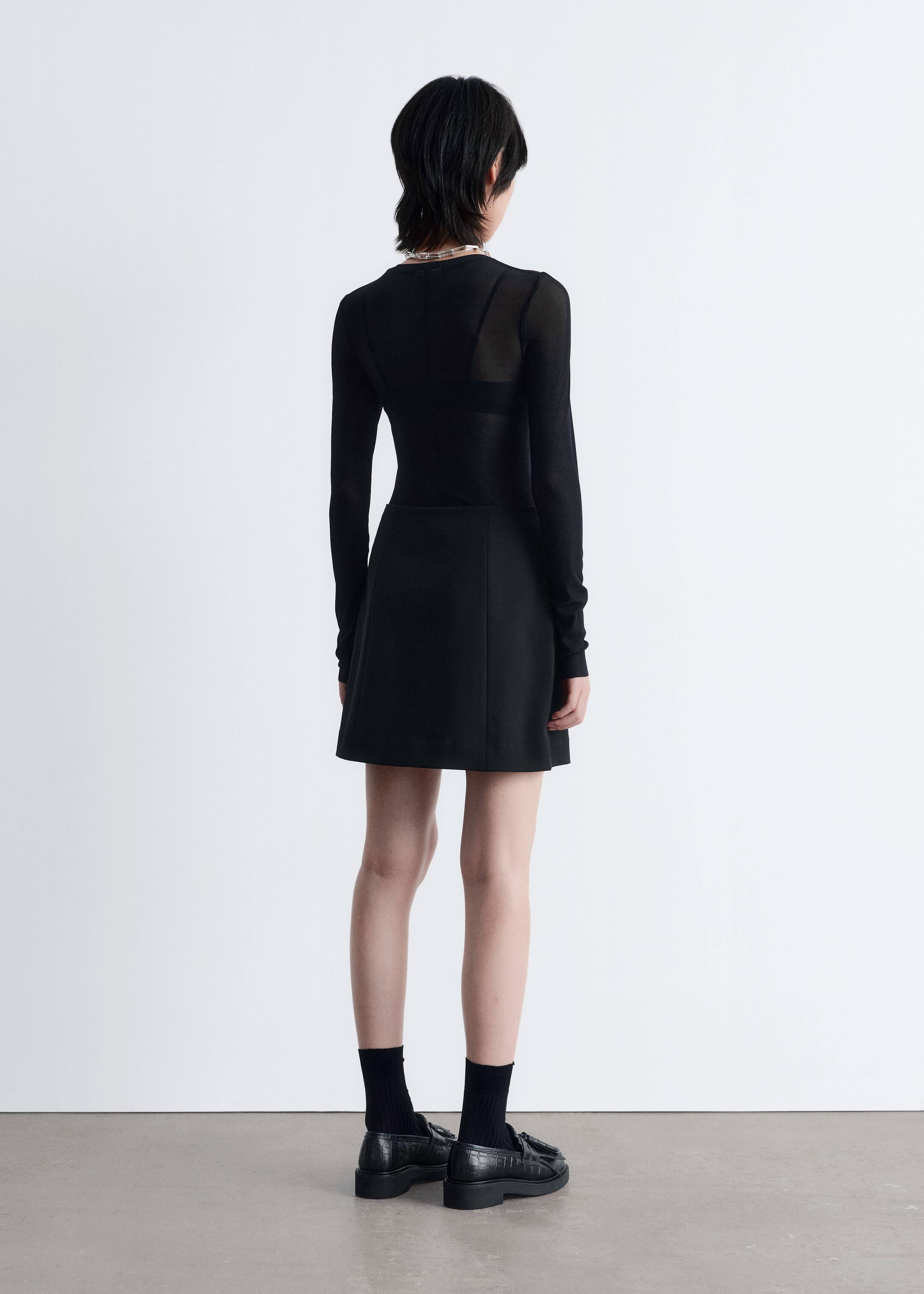Agrandir l'image: Un haut noir à manches longues, apparaissant transparent et laissant entrevoir un soutien-gorge noir en dessous, est porté avec une mini-jupe noire en ligne A, présentant des détails de coutures verticales à l'arrière. Des chaussettes noires montantes et des mocassins noirs texturés avec des glands complètent la tenue, le tout vu de dos.
