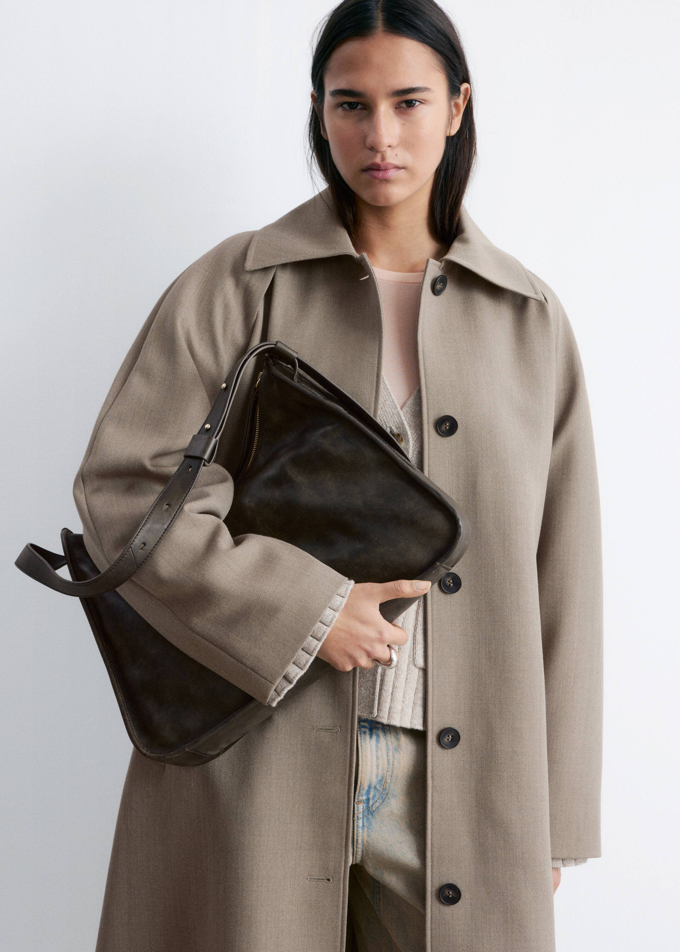 Größeres Bild anzeigen: Ein langer Mantel in Taupe wird frontal präsentiert und verfügt über einen klassischen Reverskragen, lange Ärmel sowie eine vordere Knopfleiste mit drei sichtbaren dunklen Knöpfen. Er wird offen getragen und liegt über einem hellen, gerippten Pullover, dessen Ärmelbündchen sichtbar sind.