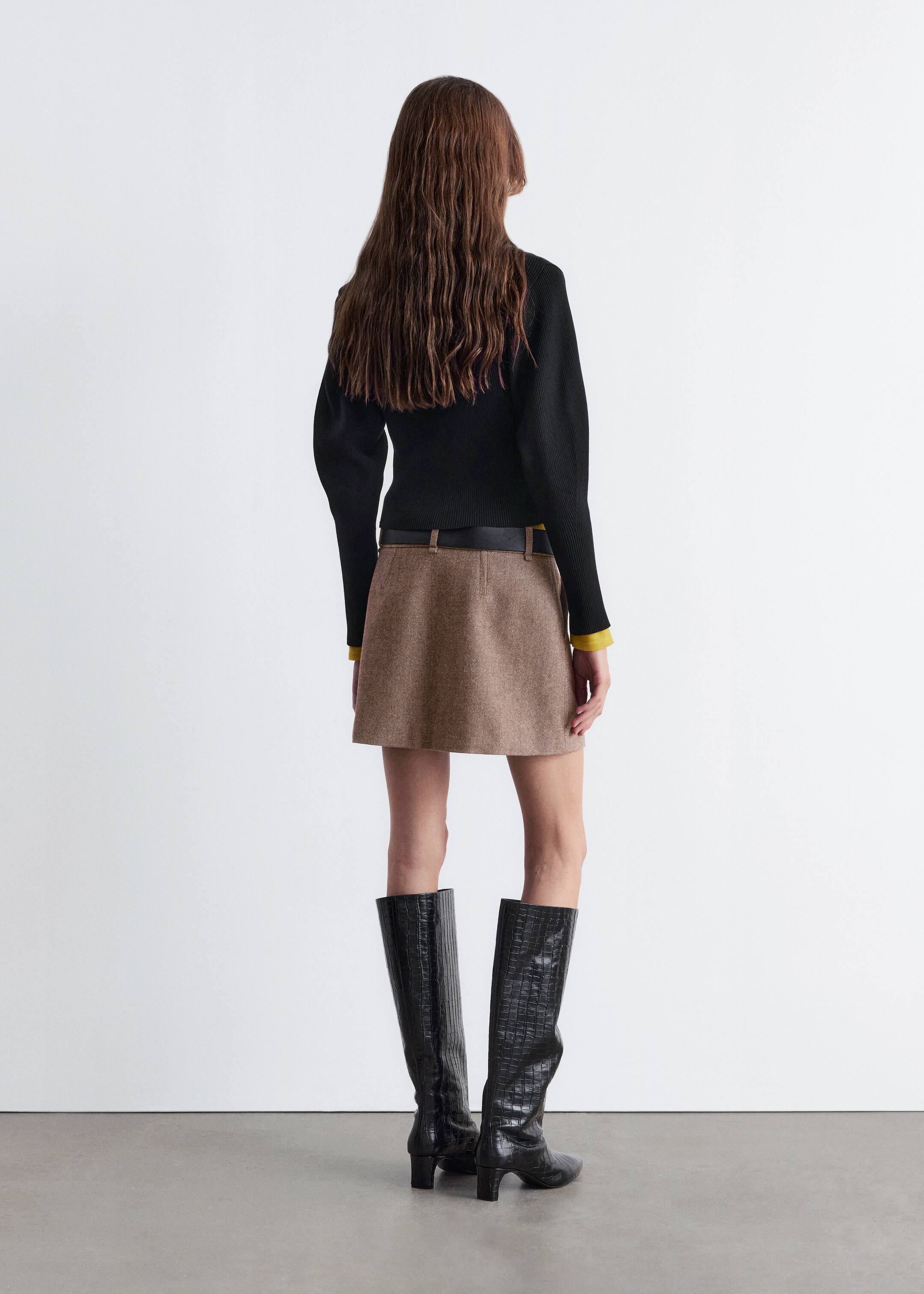 View larger image: A black long-sleeved top, featuring a ribbed texture and subtly puffed shoulders, is being worn, with a yellow cuff visible at the wrist. Below, a brown, textured mini A-line skirt is being cinched by a black belt. Black knee-high boots, displaying a crocodile-embossed pattern and block heels, are also visible, all viewed from the back.