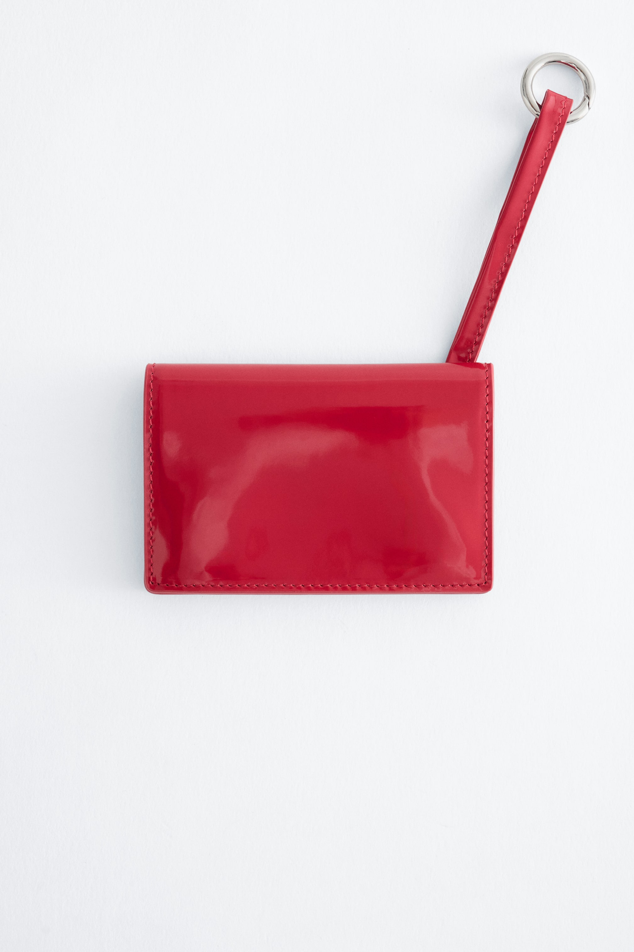 View larger image: A red patent leather card holder is shown facing forwards, featuring a glossy finish with tonal stitching along its edges. A matching red strap extends diagonally upwards from the top right corner, ending with a silver-tone metal ring.