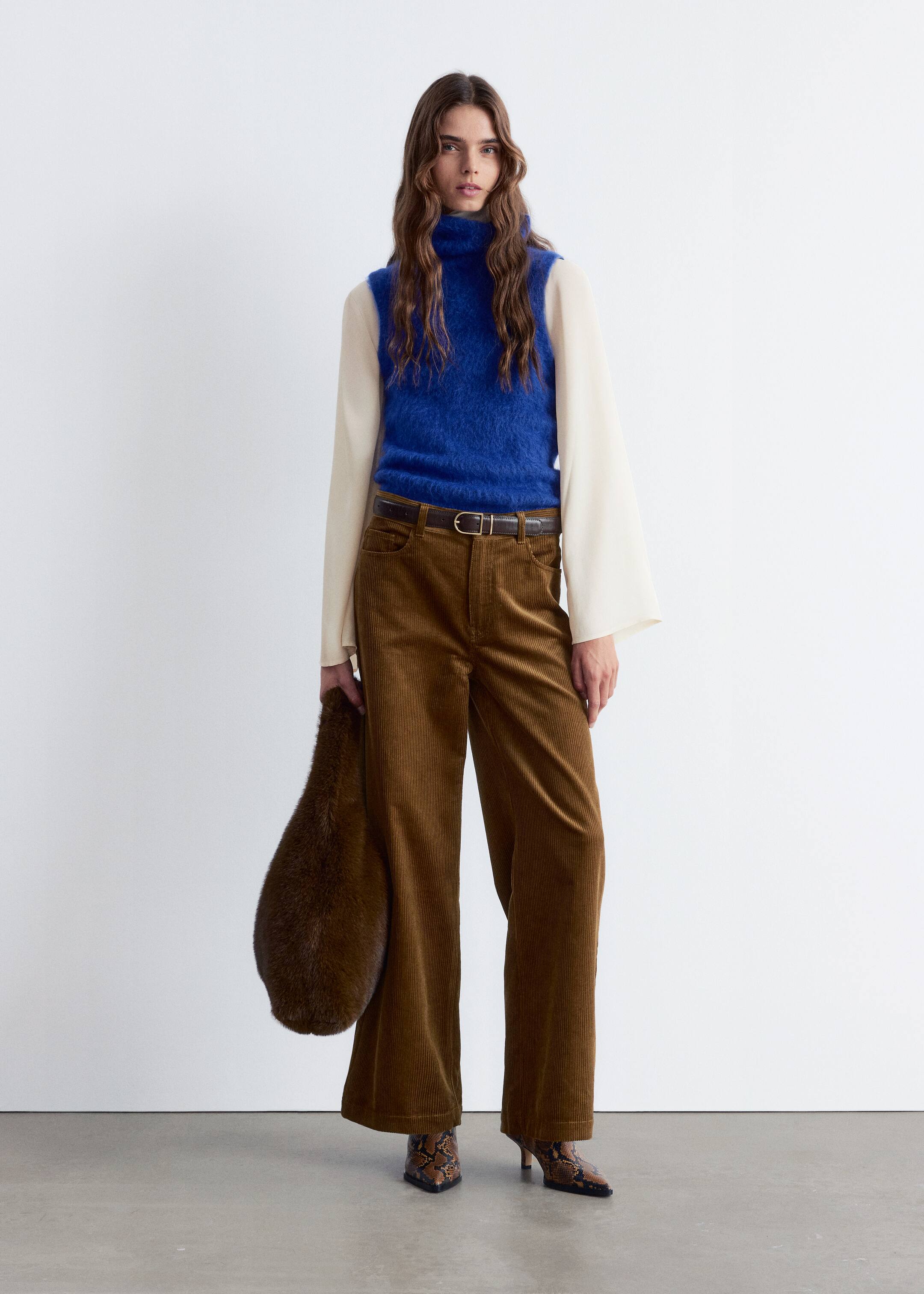 Größeres Bild anzeigen: Ein Outfit wird präsentiert, das nach vorne gerichtet ist und ein hellblaues, flauschiges ärmelloses Rollkragenoberteil zeigt, das über einer cremefarbenen langärmeligen Bluse mit weiten Glockenärmeln getragen wird. Braune Cordhosen mit weitem Bein werden getragen, mit einem dunkelbraunen Gürtel geschnürt und mit spitz zulaufenden Stiefeletten mit Schlangenhautmuster und einer dunkelbraunen länglichen flauschigen Tasche kombiniert.