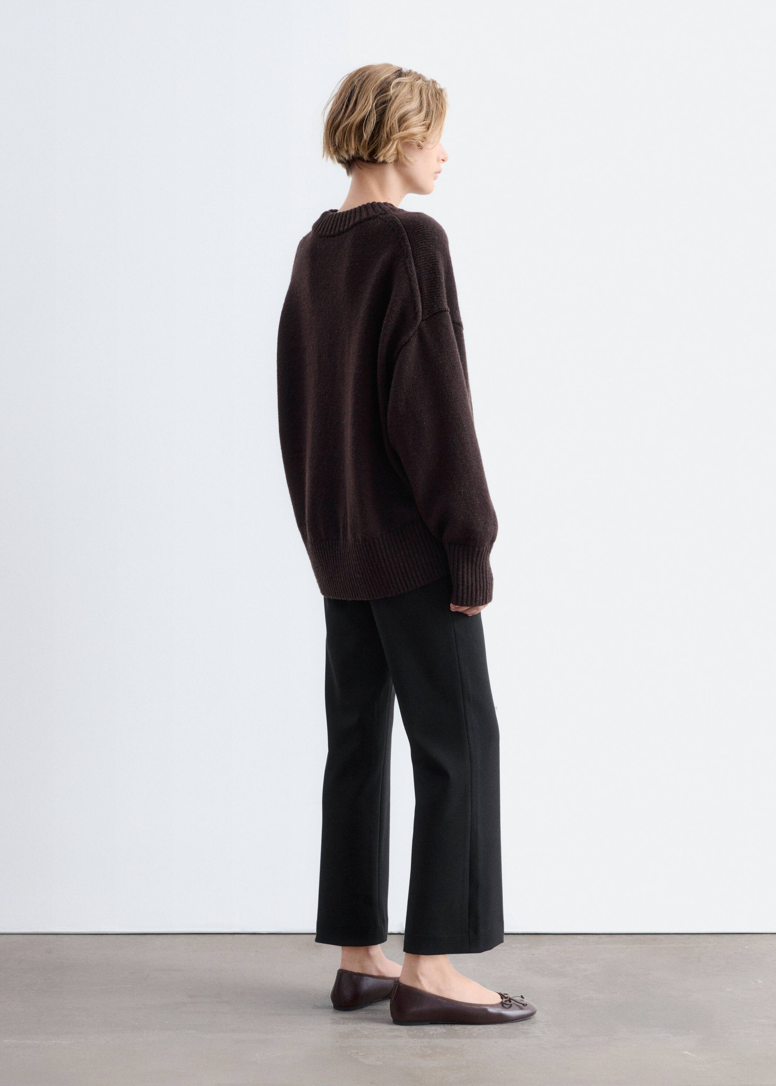 An oversized wool sweater in a dark brown knit is worn by the model in a side-back profile view. The sweater features dropped shoulders, a ribbed crewneck, and ribbed trim at the cuffs and hem, styled with black trousers and brown flat shoes.
