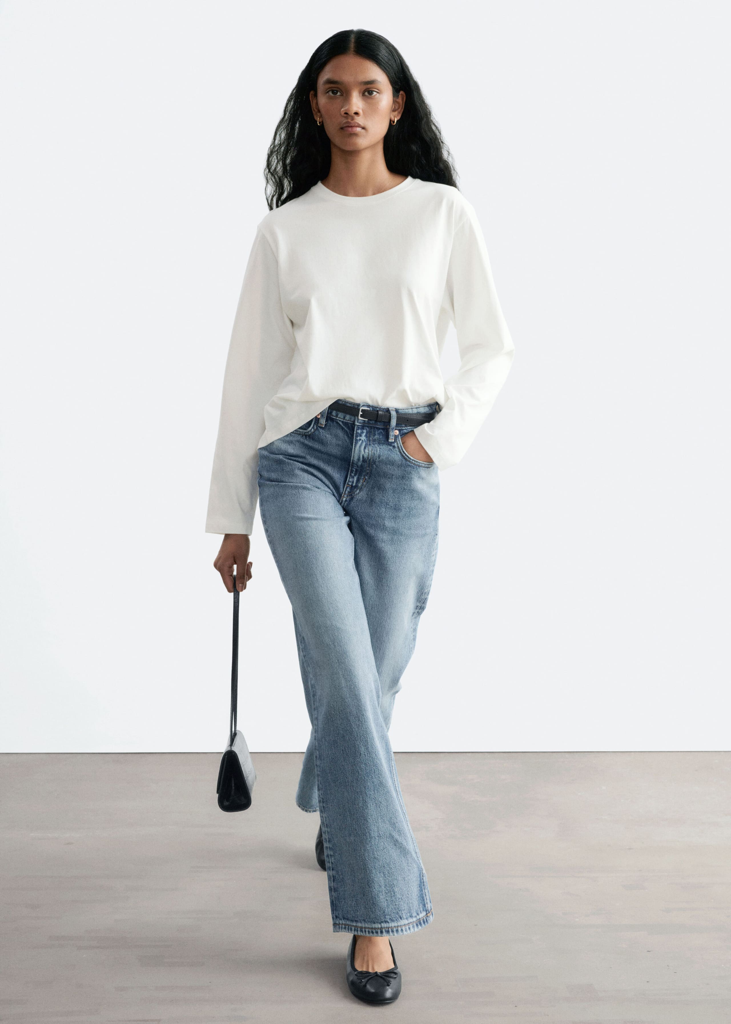 Blue straight-leg jeans with a mid-wash finish are worn by the model, who is walking forward with one hand resting in a front pocket. The jeans are styled with a thin black belt, a partially tucked-in long-sleeved white top, and black flat shoes.