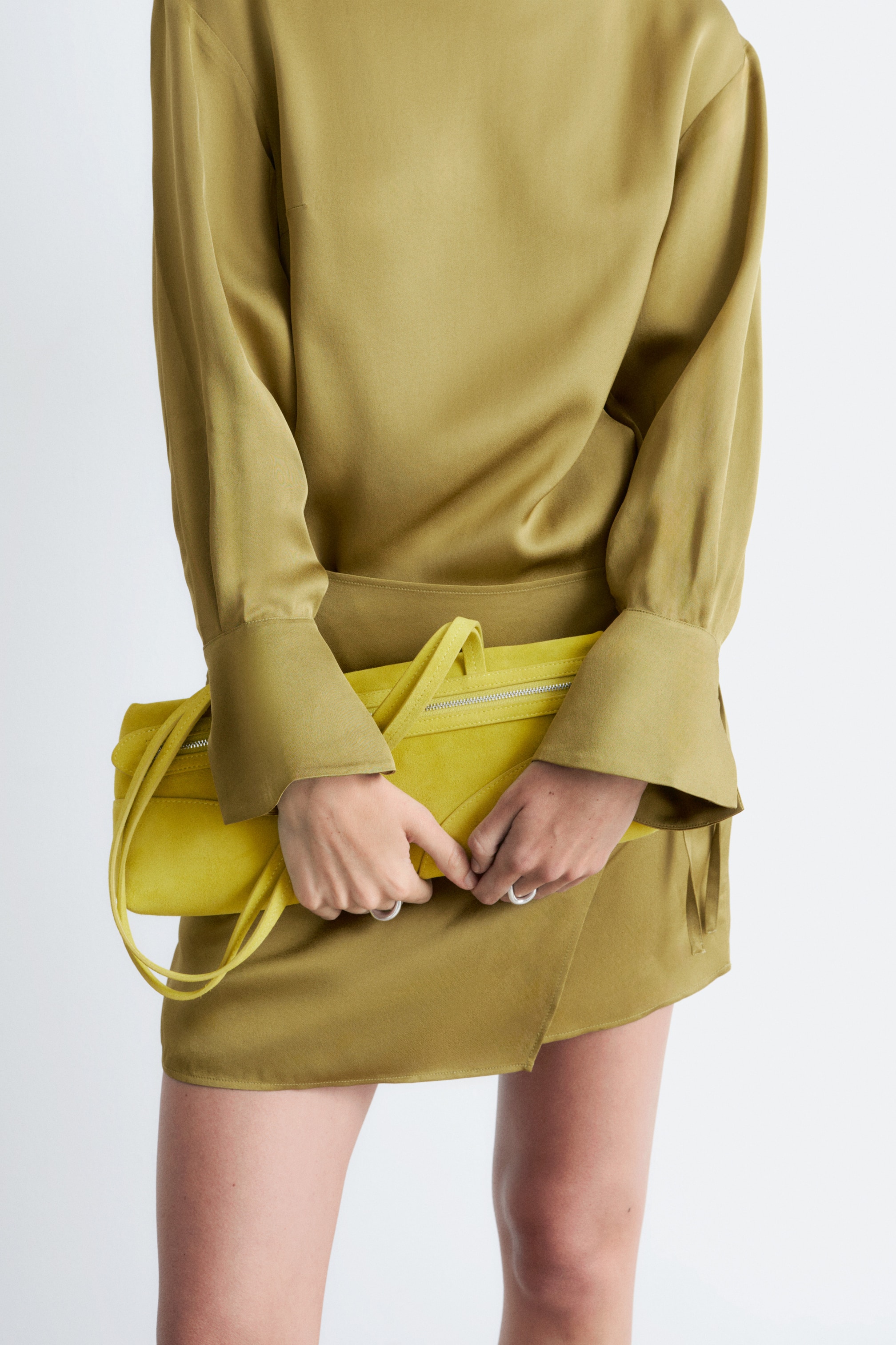 Visualizza immagine più grande: Una pochette gialla vivace con una texture simile al camoscio viene tenuta orizzontalmente da due mani, mostrando il suo design frontale. Presenta una chiusura con zip color argento lungo il bordo superiore, due sottili cinturini ad anello e un dettaglio a patta triangolare sul davanti.