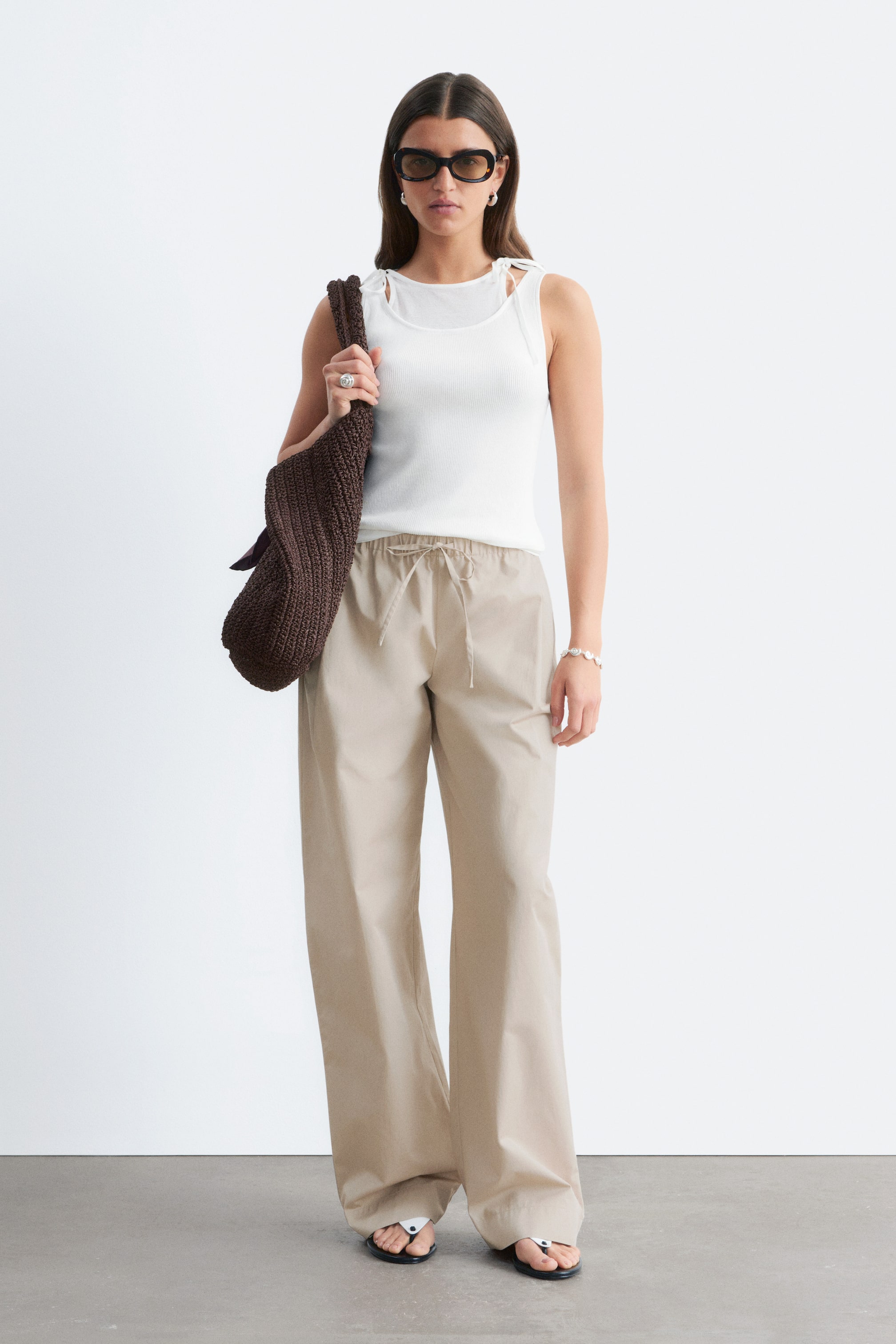 View larger image: Tan cotton twill trousers featuring a low, elasticated drawstring waist, front pleats, and slanted side pockets are worn by the model, creating a relaxed silhouette.