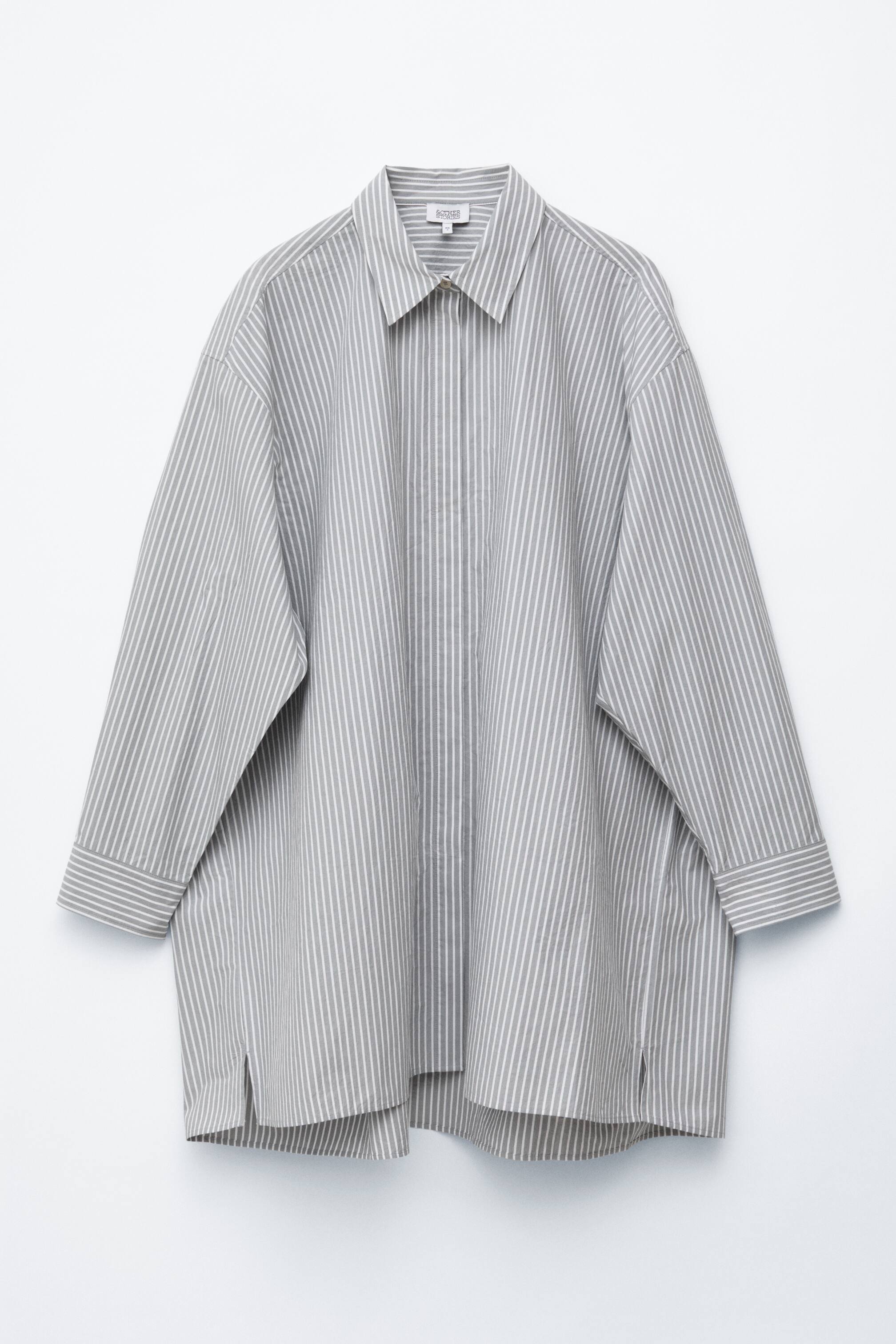 Ver imagem maior: Um vestido-camisa de riscas verticais cinza e branco, virado para a frente, exibe a sua silhueta oversized. Apresenta uma gola pontiaguda, mangas compridas com punhos, carcela de botões oculta e delicadas aberturas laterais na bainha.