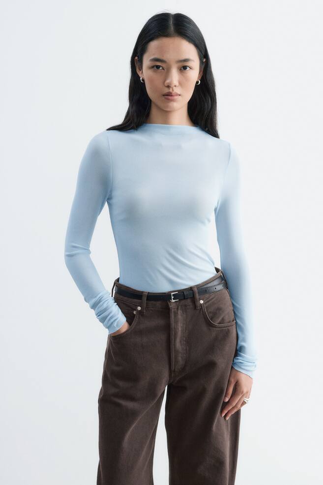 A light blue, long-sleeved ribbed silk top with a high neckline is being worn by the model. The fitted garment is tucked into dark brown trousers secured with a slim black belt. - 1
