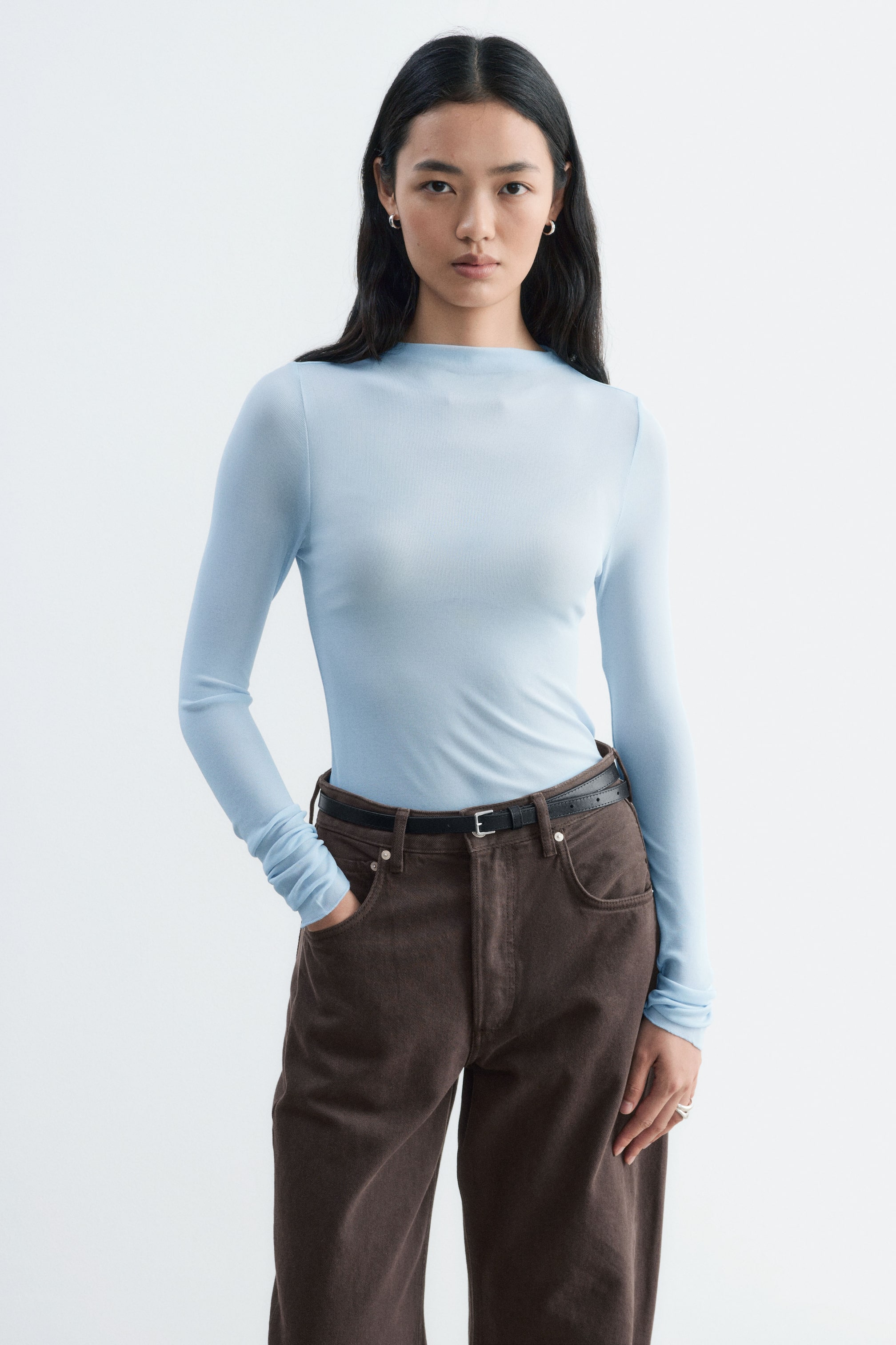 Vezi imaginea mai mare: Un top din mătase cu nervuri, bleu deschis, cu mâneci lungi și decolteu înalt, este purtat de model. Piesa ajustată este introdusă în pantaloni maro închis, strânși cu o curea subțire neagră.
