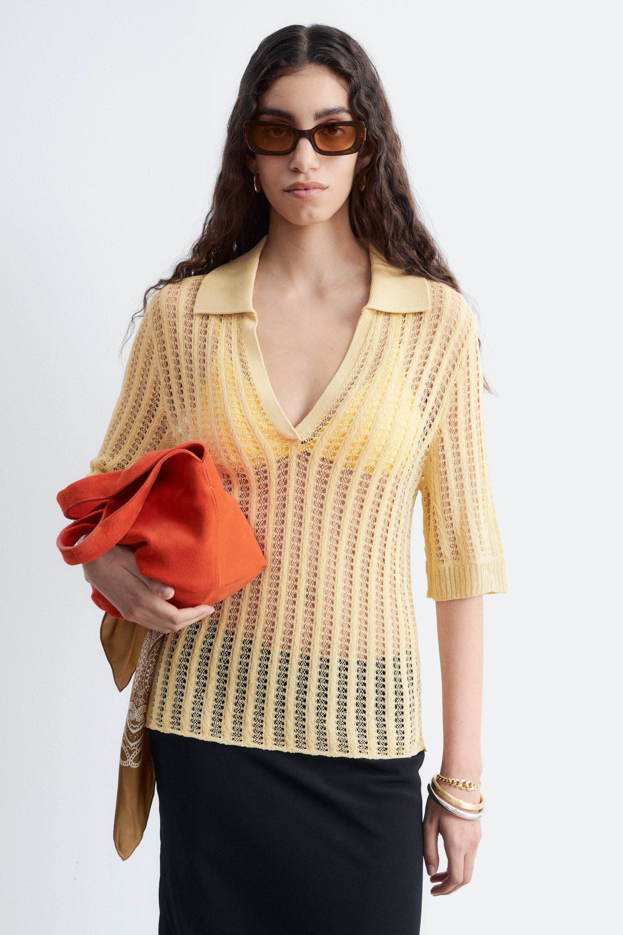 Agrandir l'image: Le polo en maille ajourée jaune clair présente un col à revers, un décolleté en V et des manches mi-longues avec des poignets côtelés. Il affiche une texture transparente à rayures verticales, superposée à un sous-vêtement jaune vif et une jupe noire.