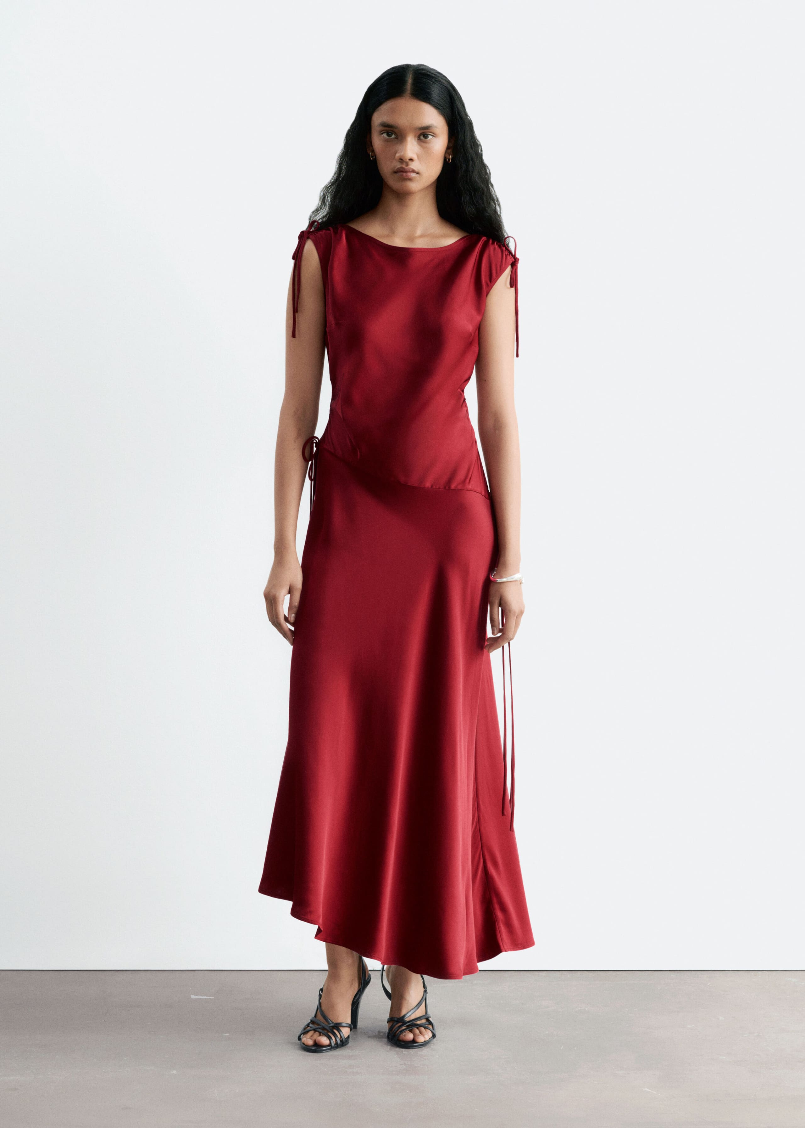 The model is wearing a red satin midi dress featuring drawstring shoulders and an asymmetric dropped waist. The dress has a draped silhouette with an asymmetric hemline and is paired with black strappy heeled sandals.