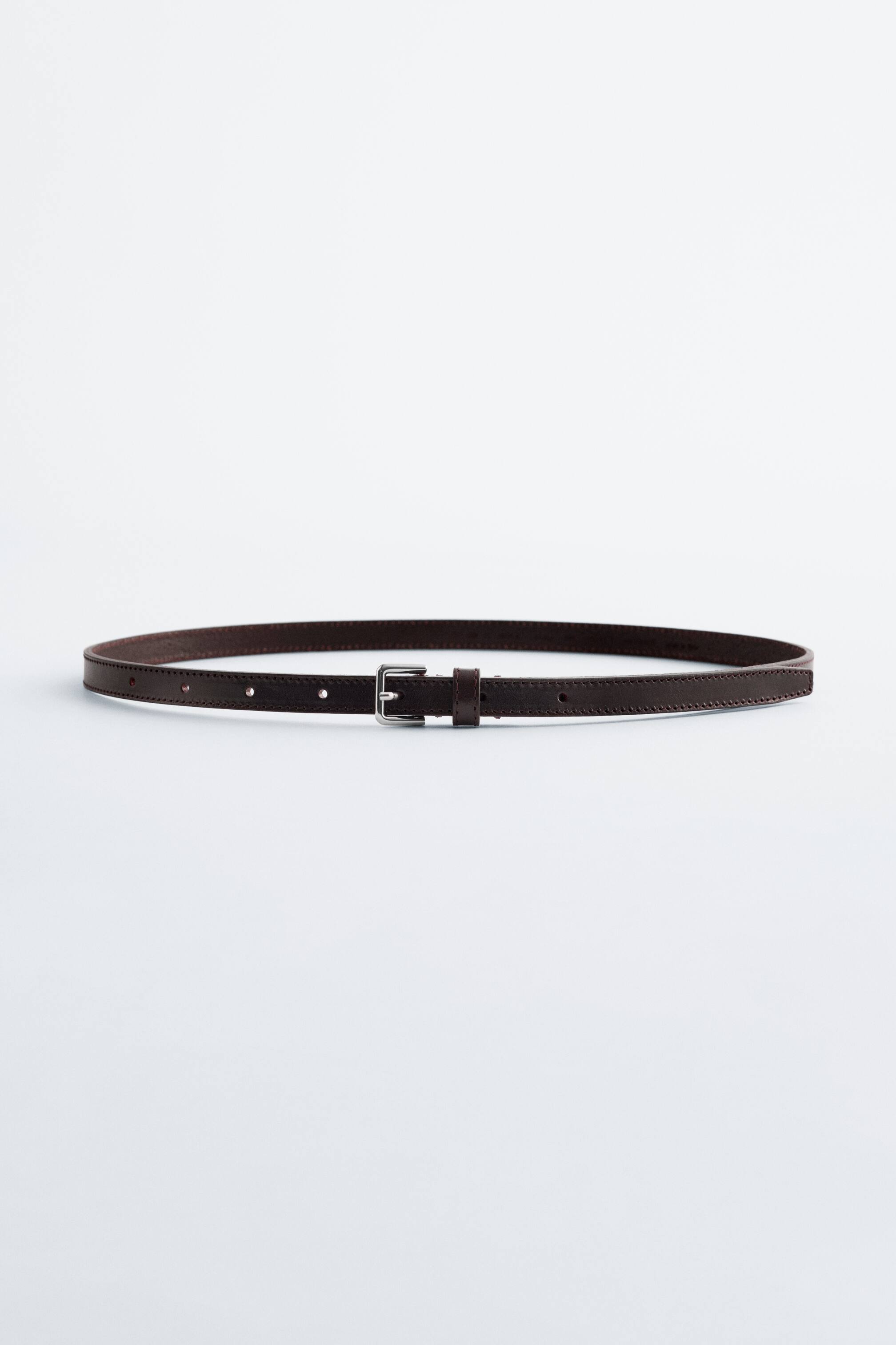 Näytä suurempi kuva: Tumma ruskea, pehmeä nahkavyö makaa tasaisesti etupuolella, ja siinä on hopeasävyinen neliönmuotoinen solki ja kulmikas kärki. Vyössä on ohut hihna, jossa on hienovaraiset reunatikkaukset ja useita säätöreikiä.