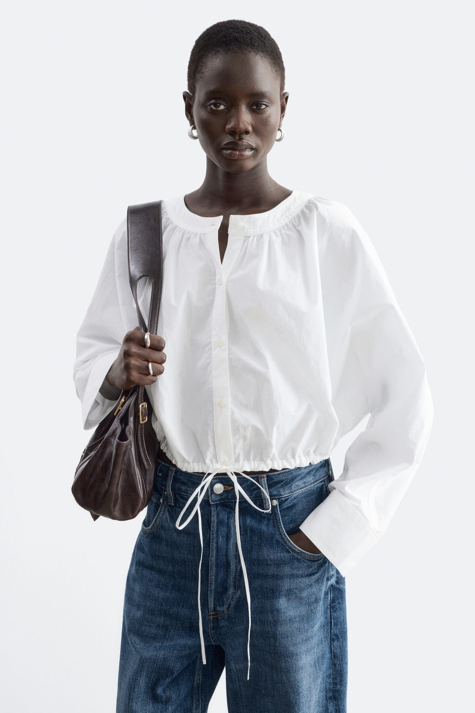 Een witte cropped katoenen blouse met een geplooide halslijn, een knoopsluiting aan de voorkant en een verstelbare zoom met trekkoord en lange strikceintuur wordt gedragen door het model. De blouse wordt gecombineerd met een blauwe denim jeans, en het model draagt een donkerbruine leren schoudertas. - 1