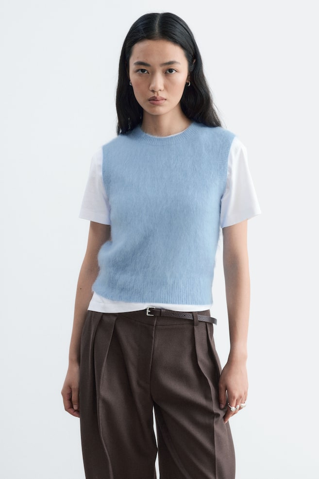 Een lichtblauwe gebreide vest van mohairmix met ronde hals en een zachte, pluizige textuur wordt gedragen door het model. Het vest is gelaagd over een wit T-shirt met korte mouwen en gestyled met bruine geplooide broek. - 1