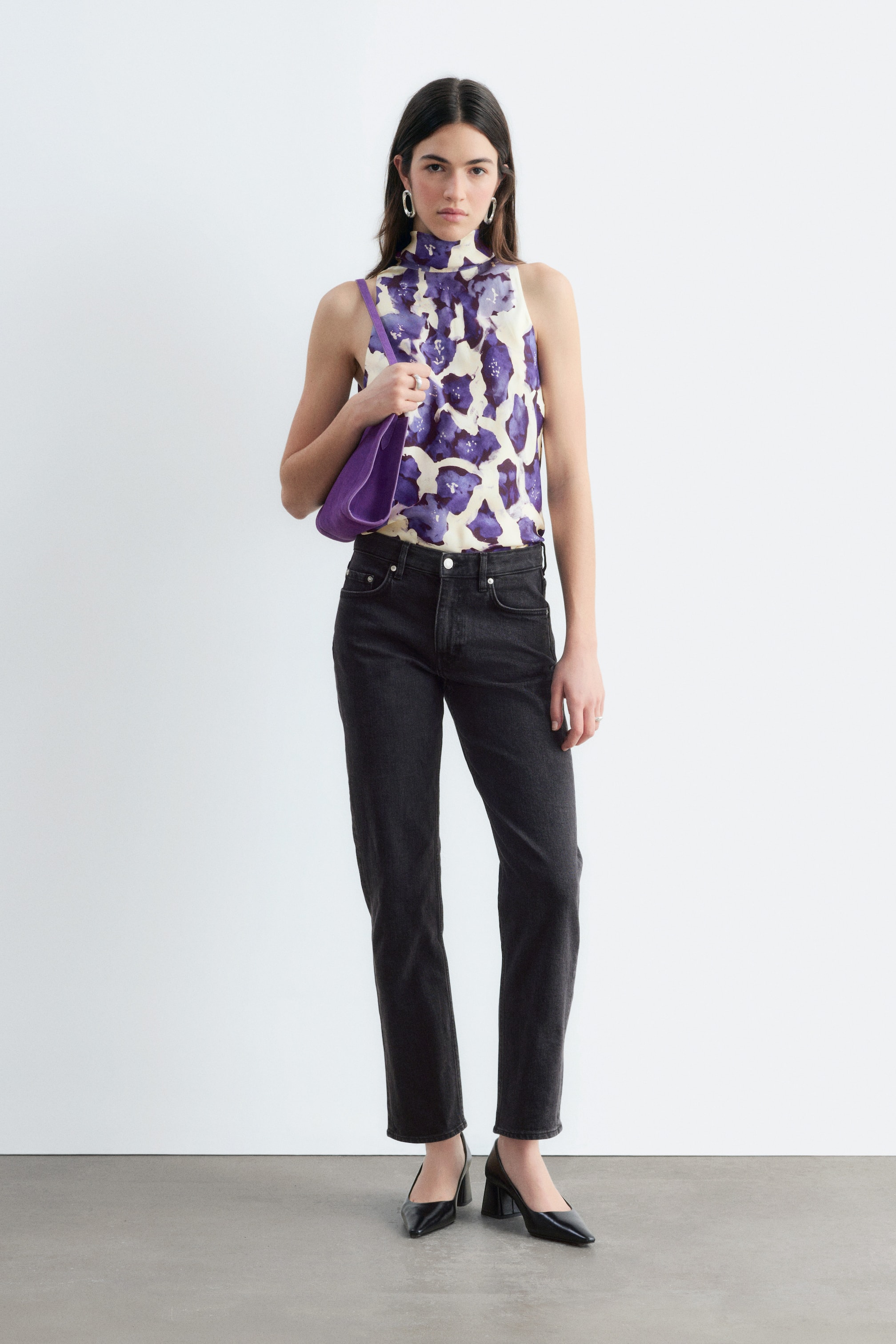 Agrandir l'image: Le mannequin porte un jean noir à cinq poches, taille normale, avec une jambe slim arrivant à la cheville. Le jean est associé à un haut sans manches à motif violet et crème, rentré à l’intérieur, ainsi qu’à des escarpins noirs à talon bloc.