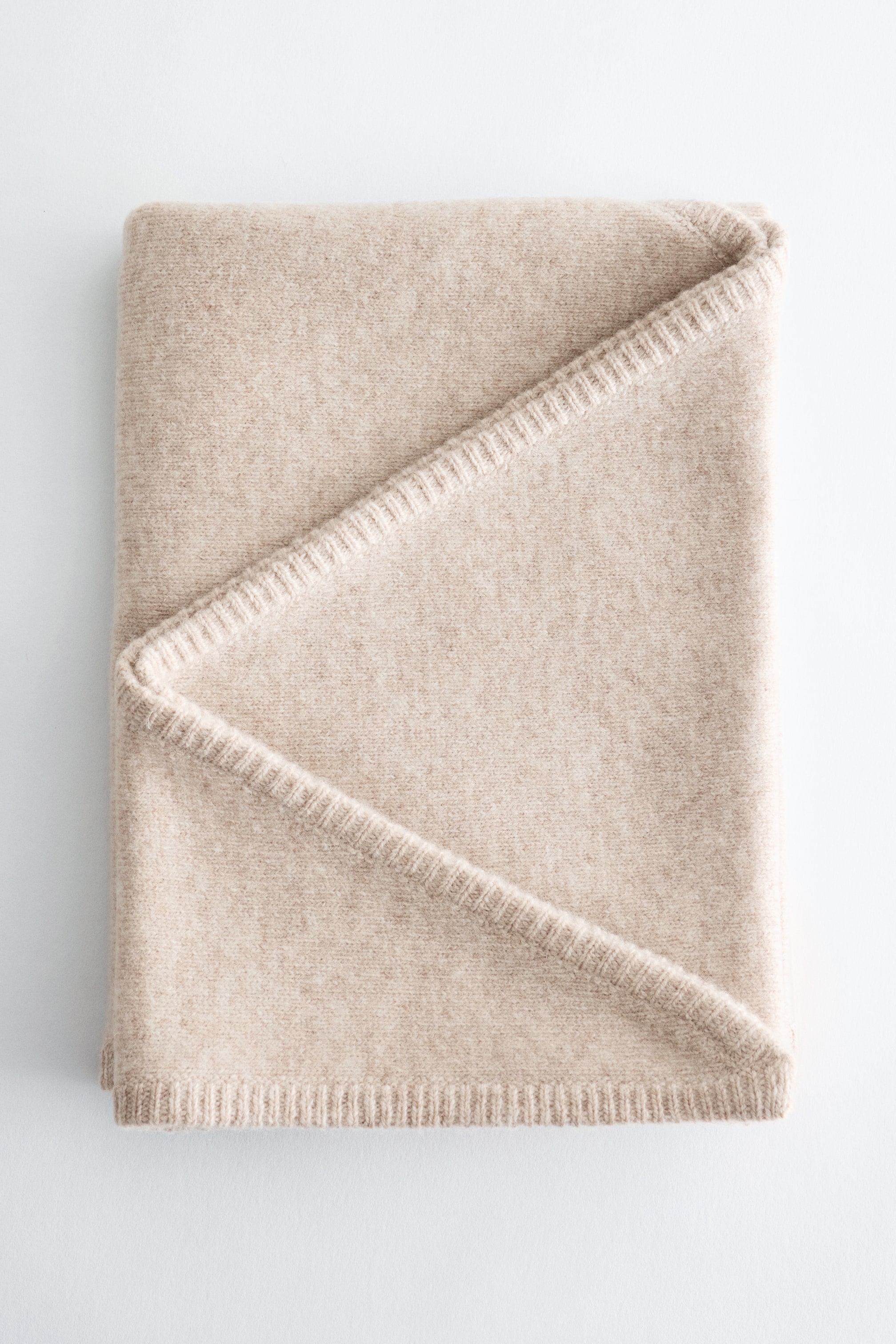 Agrandir l'image: Un textile beige clair est posé à plat, plié en forme rectangulaire. Un pli diagonal traverse sa surface, révélant une finition en maille côtelée ton sur ton le long des bords.