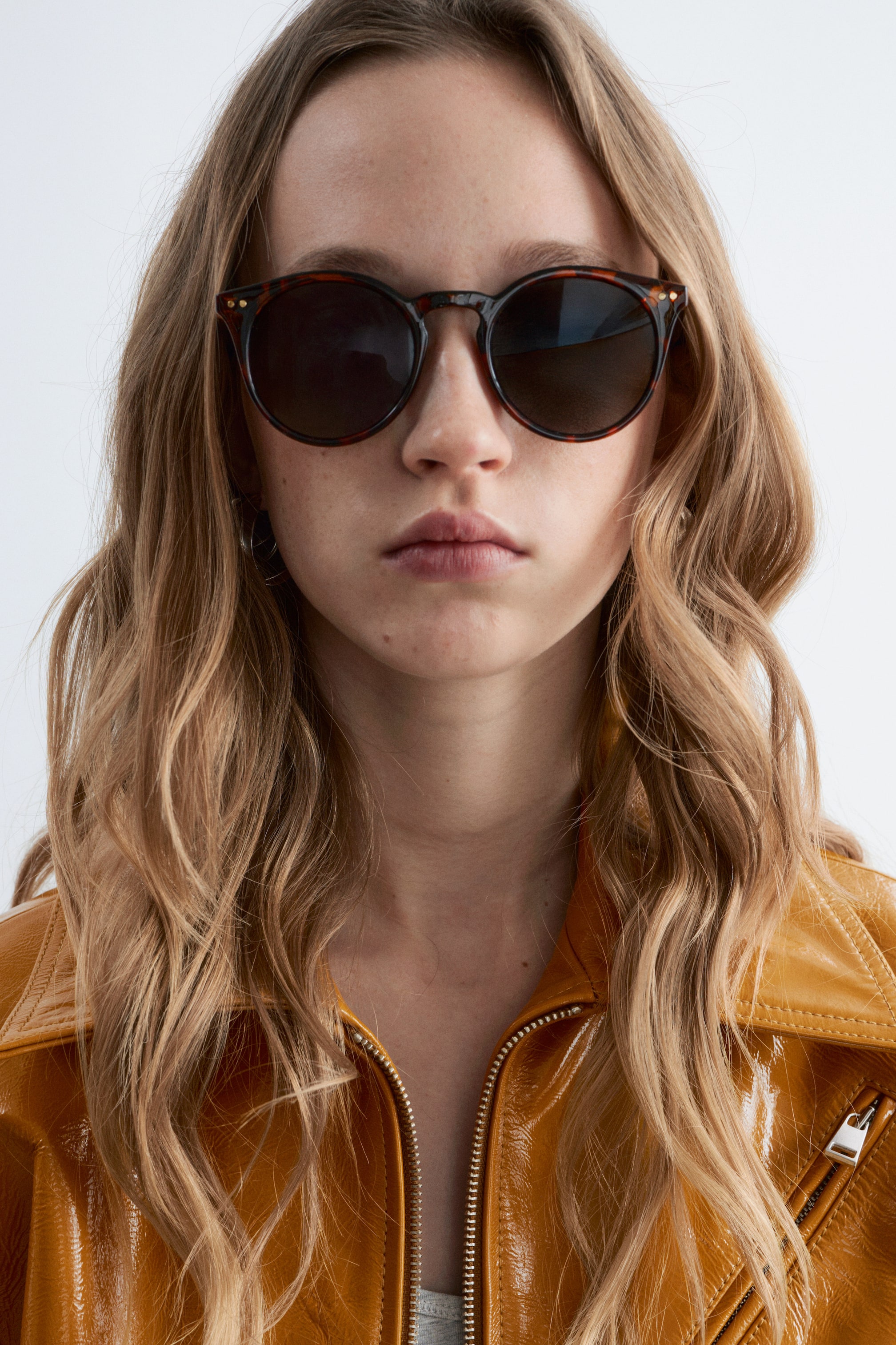 Agrandir l'image: Les lunettes de soleil présentent des montures rondes en écaille de tortue avec des verres teintés foncés. De petits clous dorés ornent le devant de la monture près des charnières, et elles sont portées face vers l'avant.