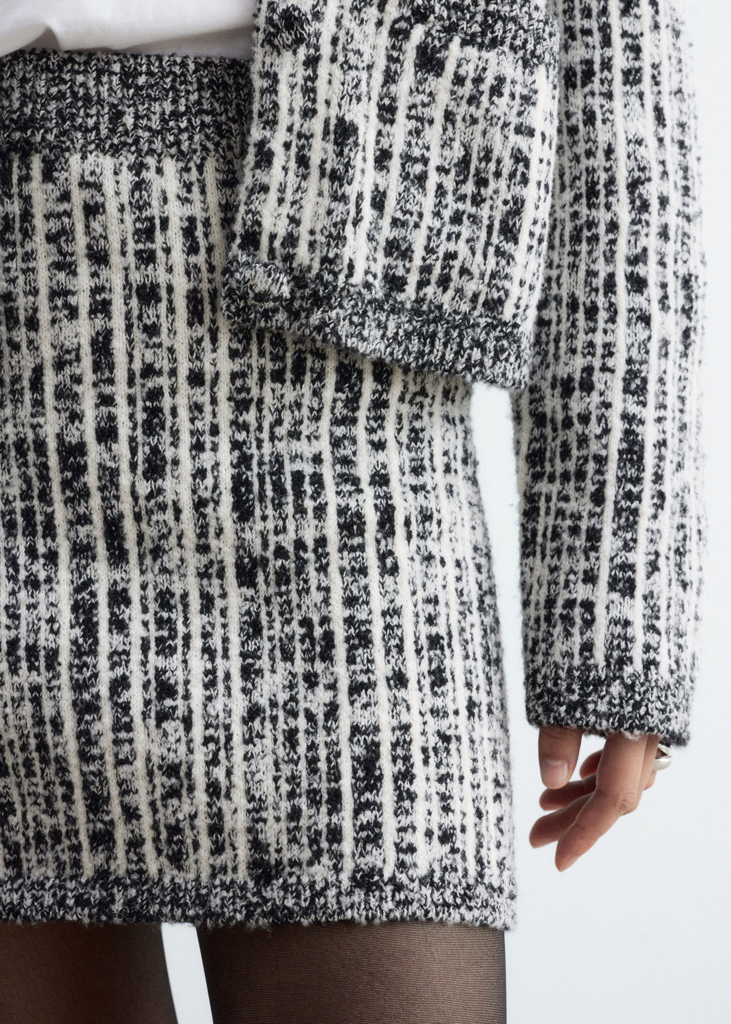 The model is wearing a matching black-and-white knit skirt and jacket with a textured vertical pattern. The coordinated set is styled with a white top and sheer black tights.