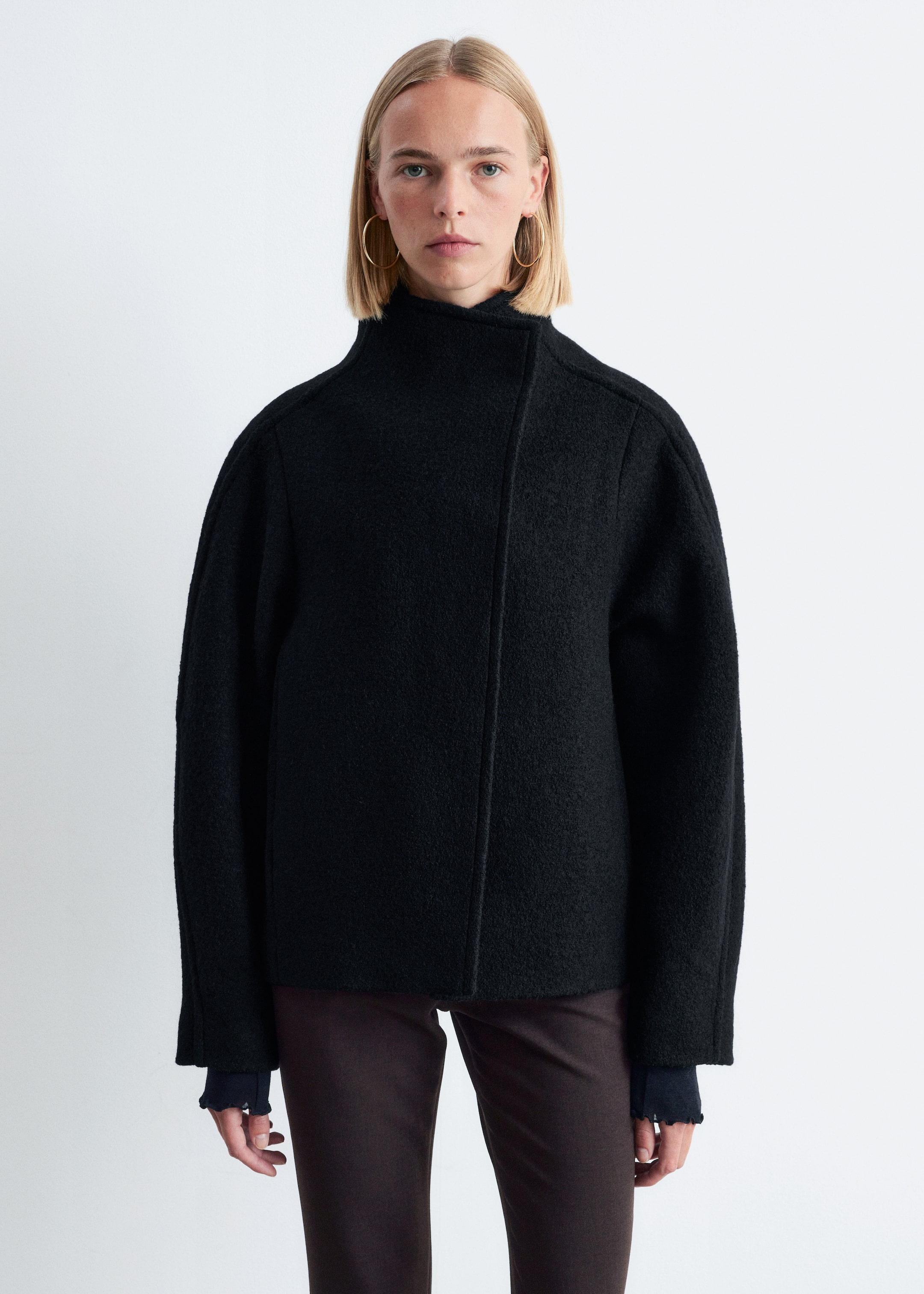 View larger image: A black jacket is being shown, featuring a textured fabric, a high stand-up collar, and an asymmetrical front closure. The jacket is designed with dropped shoulders and wide sleeves, presenting a relaxed, cropped fit, with a dark blue ruffled cuff being visible underneath the sleeves.