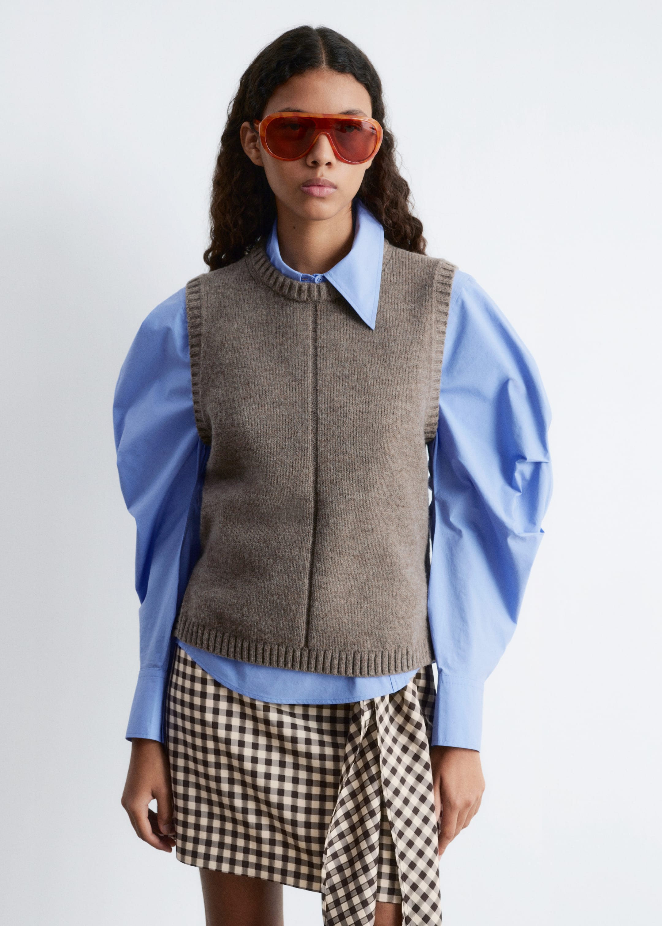 Agrandir l'image: Un gilet en tricot marron, avec un col rond, une bordure côtelée et une couture frontale verticale, est superposé à une chemise à col bleu clair avec des manches bouffantes volumineuses. Une mini-jupe à carreaux marron et crème avec un détail asymétrique enveloppant complète la tenue.