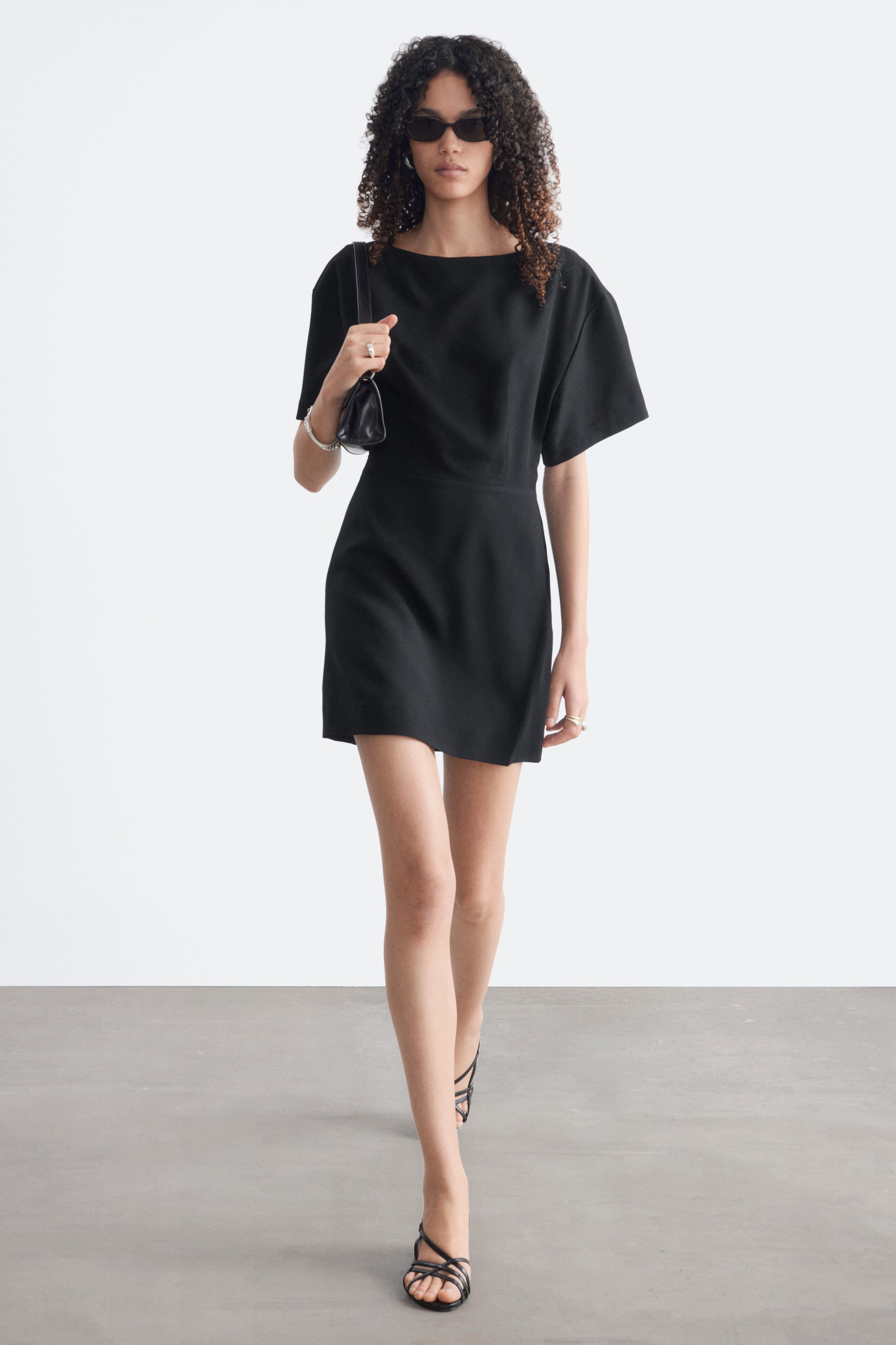 View larger image: A black mini dress features a wide boat neckline and short, loose sleeves. It showcases a defined waistline leading to a straight skirt that ends above the knee.