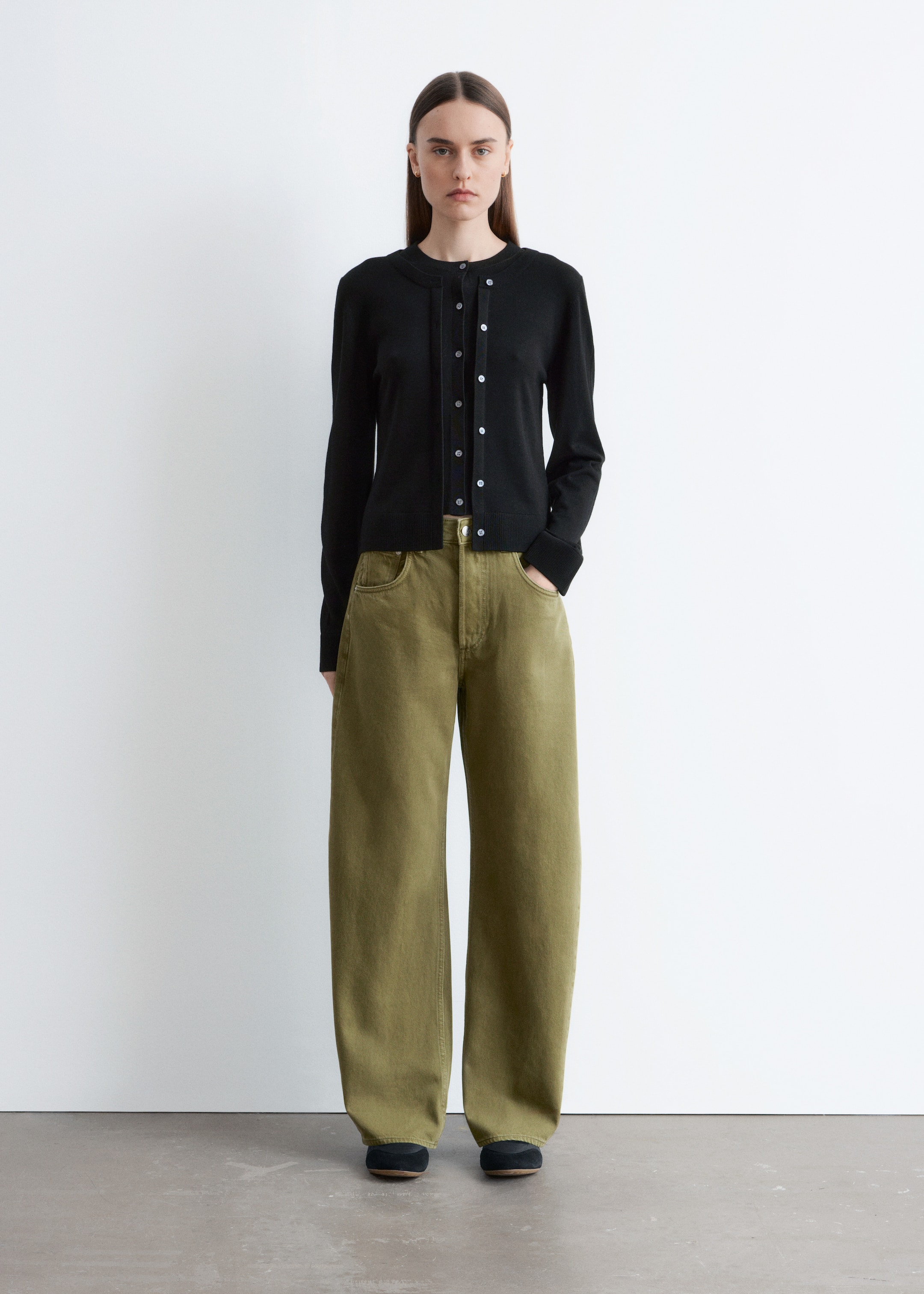 Ver imagem maior: Um cardigã preto de decote redondo é apresentado de frente, exibindo um fecho frontal com botões em tom claro, mangas compridas com punhos canelados e uma barra canelada. Está a ser coordenado com calças wide-leg verde-oliva.