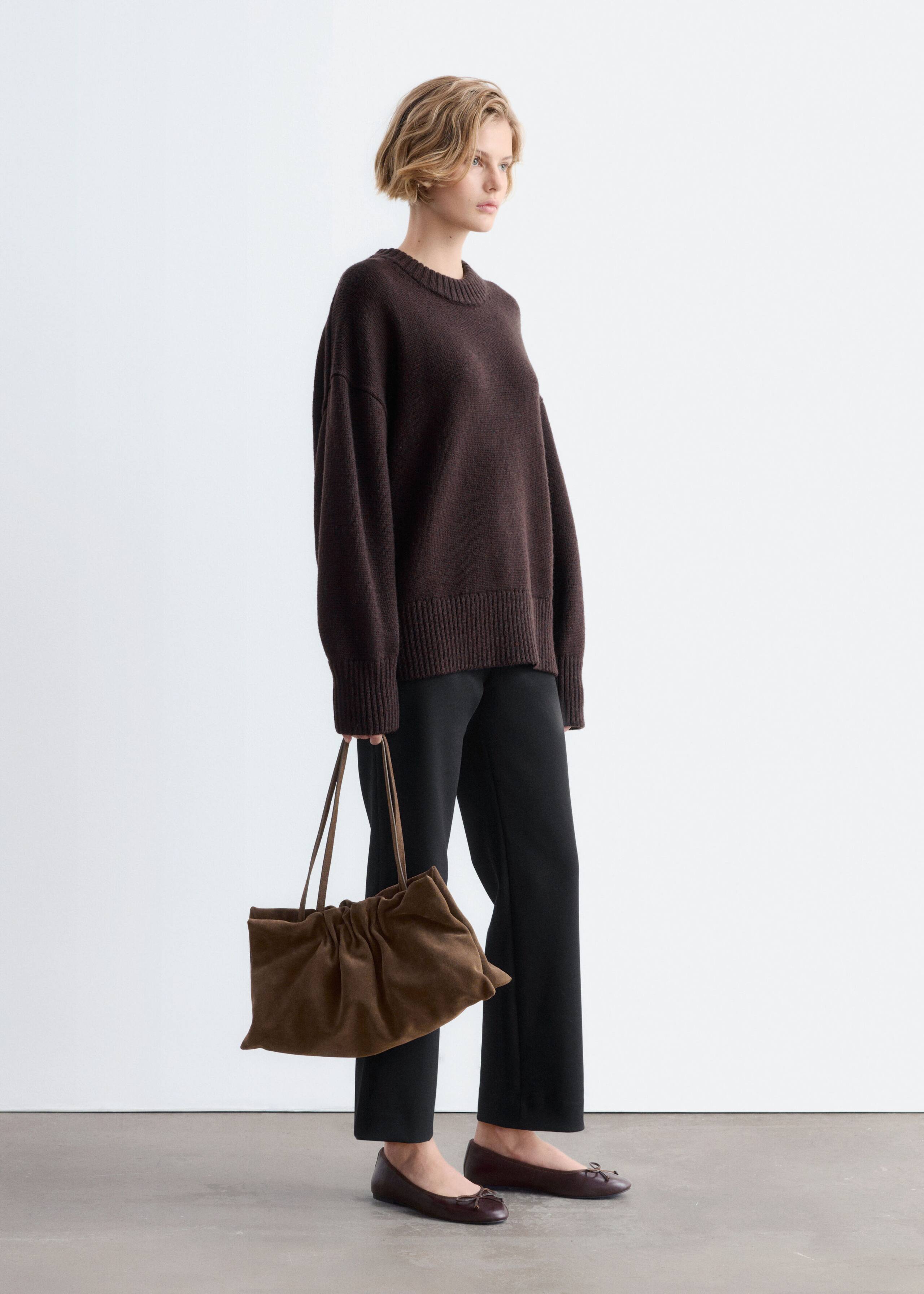 The model is wearing an oversized dark brown wool jumper featuring a crew neck, dropped shoulders, and ribbed trim at the cuffs and hem. It is styled with black straight-leg trousers, flat brown shoes, and a brown suede handbag held by the model.